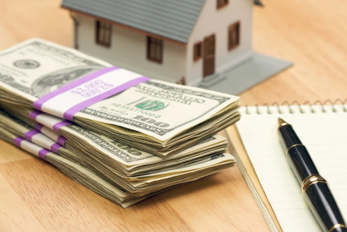 A stack of money next to a model house and a pen