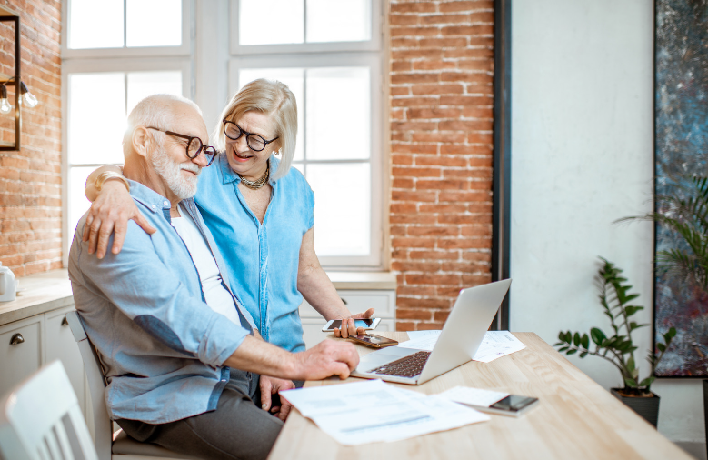 An elderly couple is sitting at a table looking at a laptop.