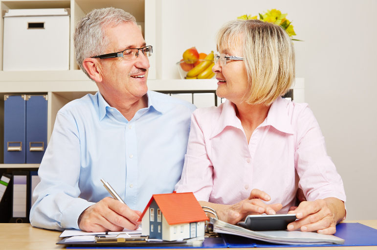 An elderly couple is sitting at a table with a model house and a calculator.
