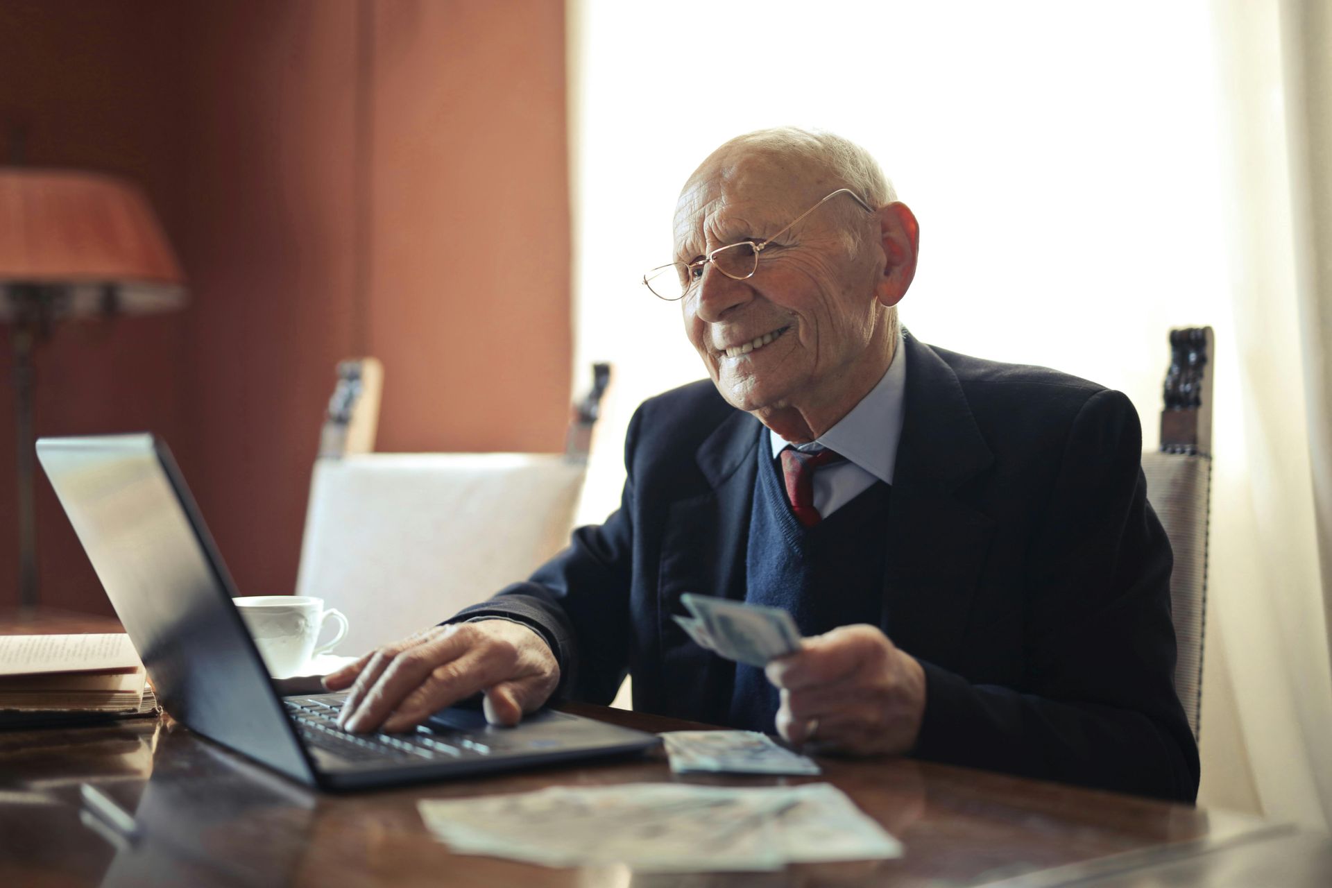 An elderly man is sitting at a table using a laptop computer and holding money.