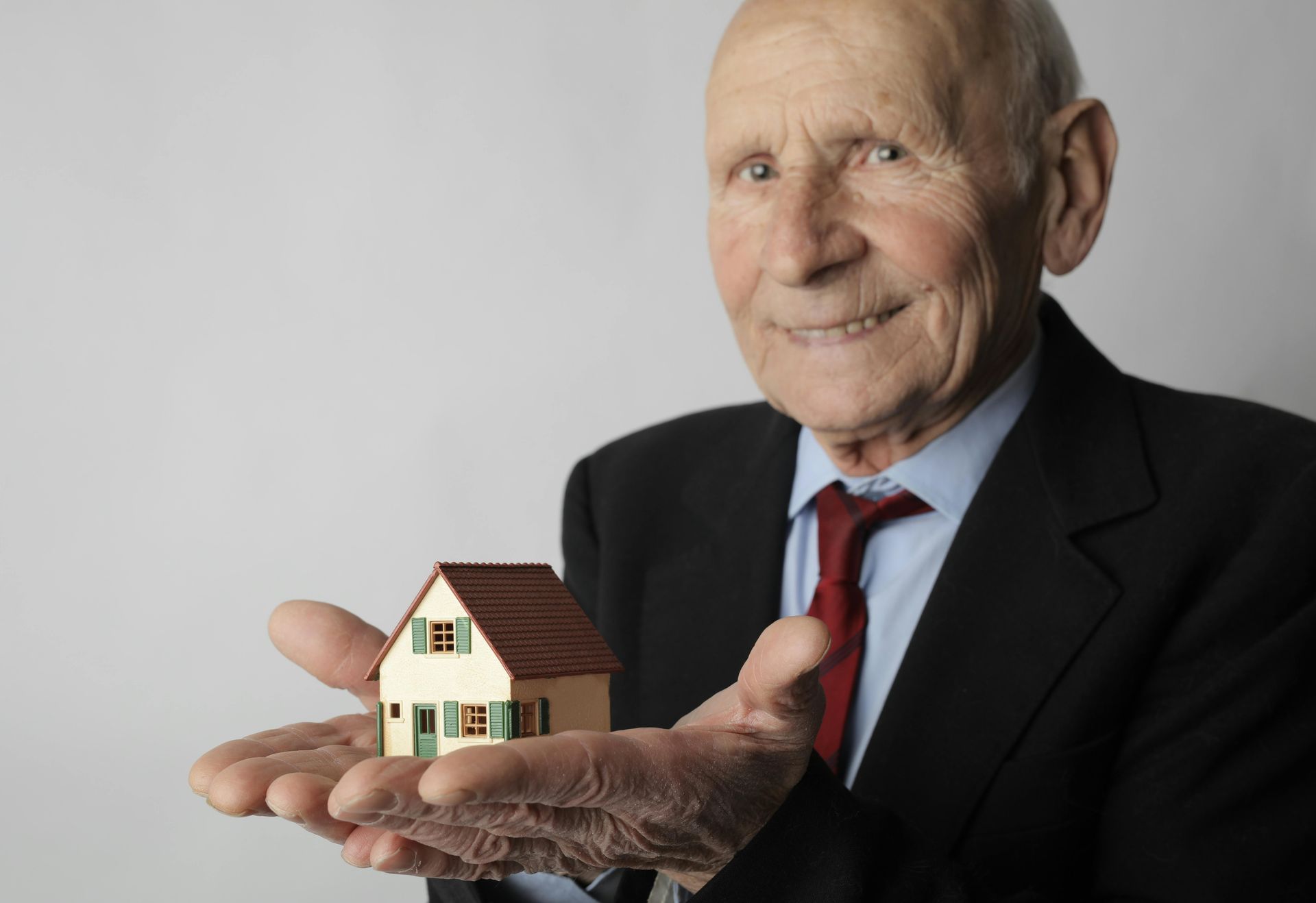 An elderly man in a suit and tie is holding a small house in his hands.