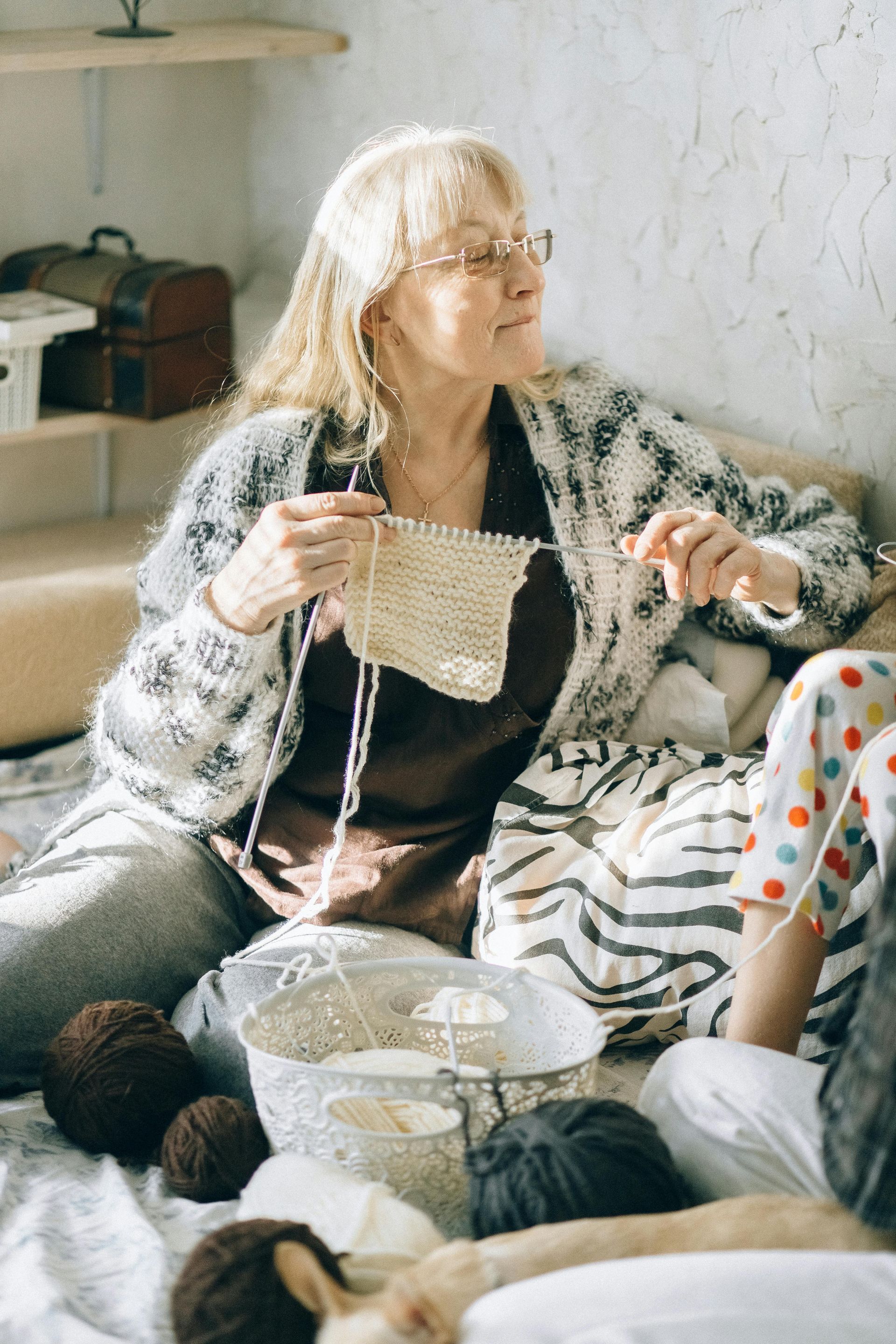 An elderly woman is knitting while sitting on a couch.