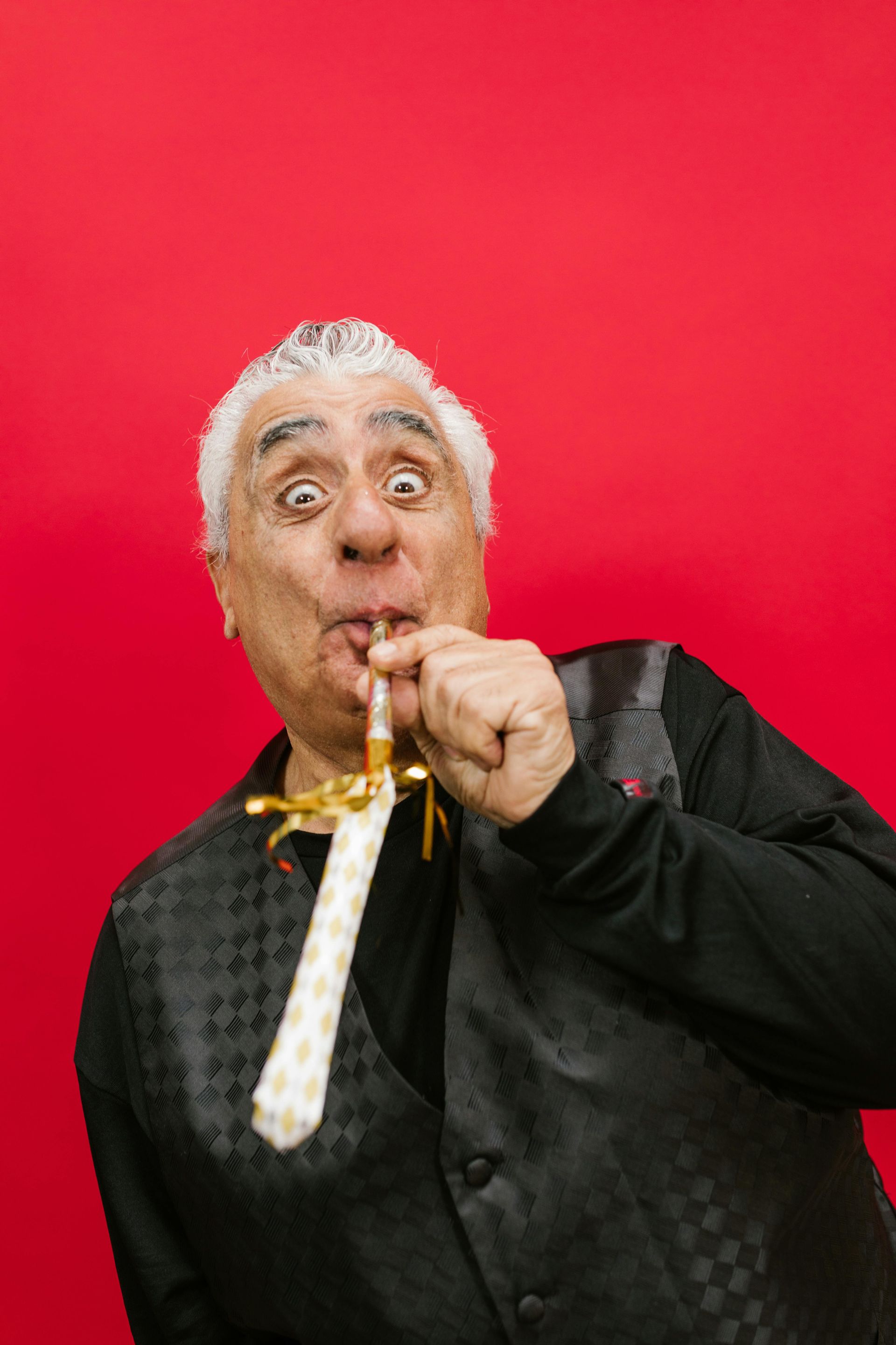 A man in a black vest and tie is blowing a party horn.