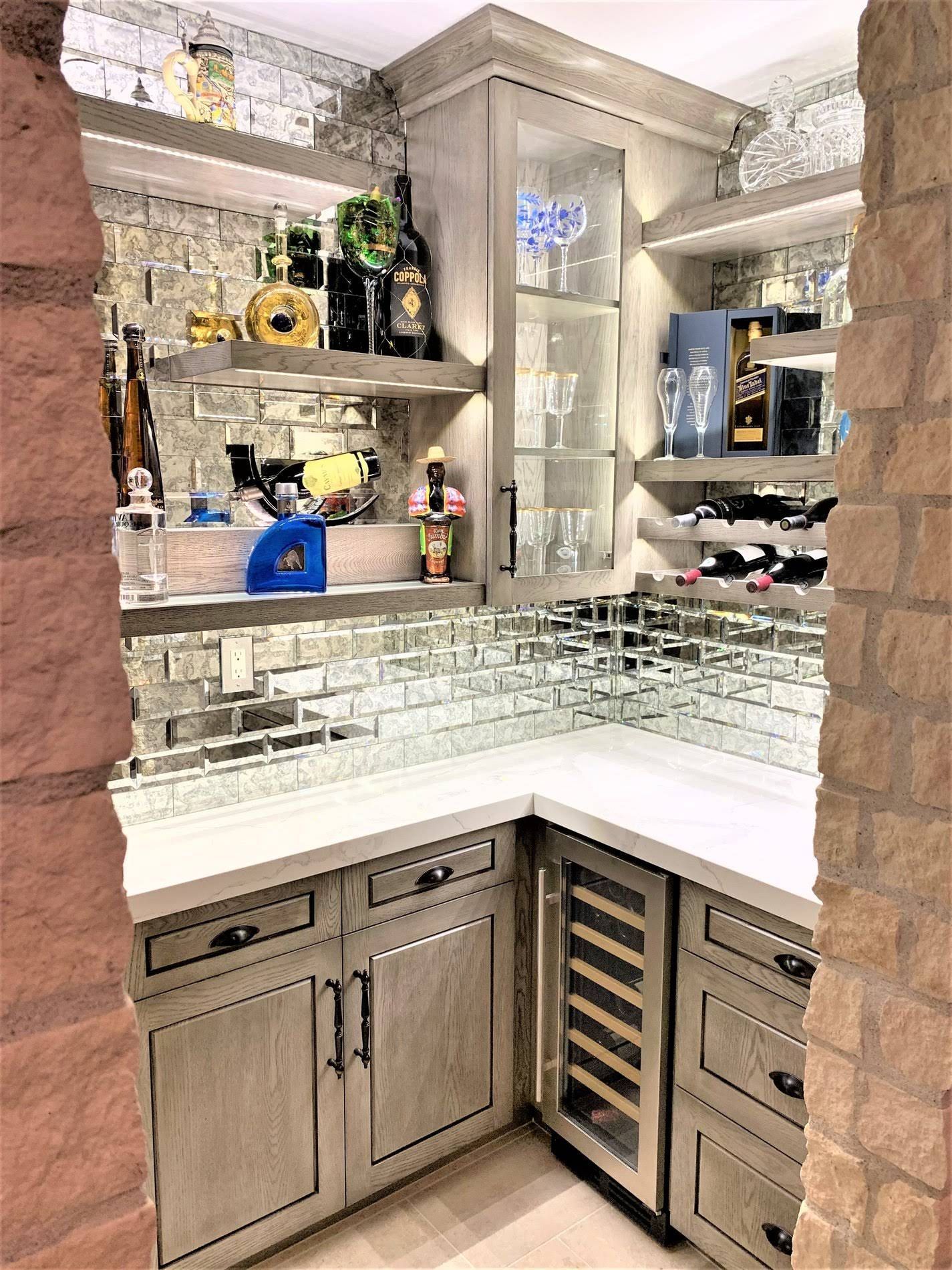 Corner home bar with gray cabinets, mirrored backsplash, shelves of liquor and glassware.
