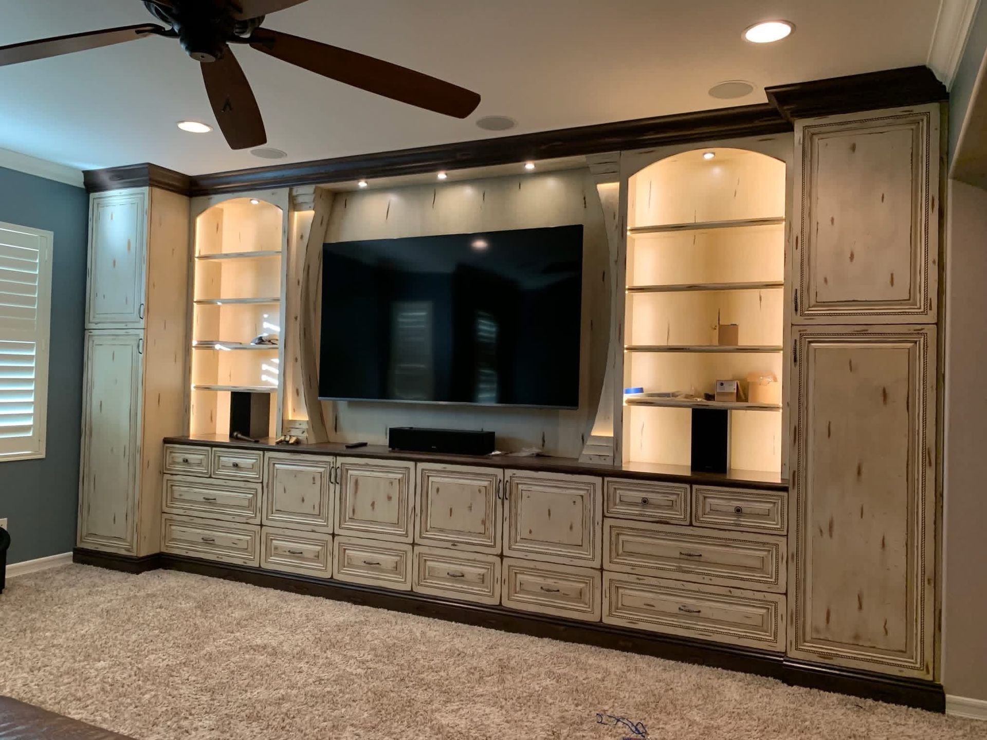 Custom built-in entertainment center with TV, shelves, and cabinets. Light beige with dark brown trim and lighting.