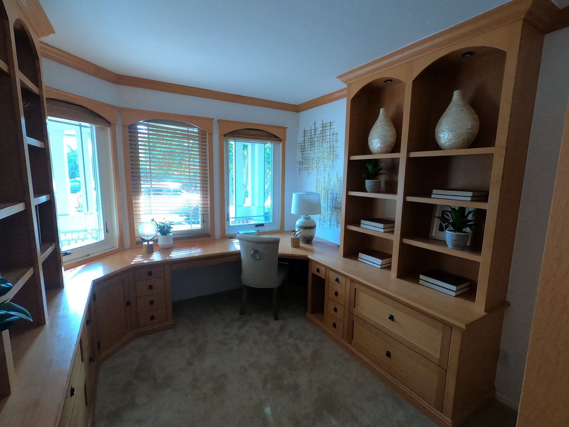 A home office with a built-in desk, shelves, and a window. Light wood furniture and neutral carpet.