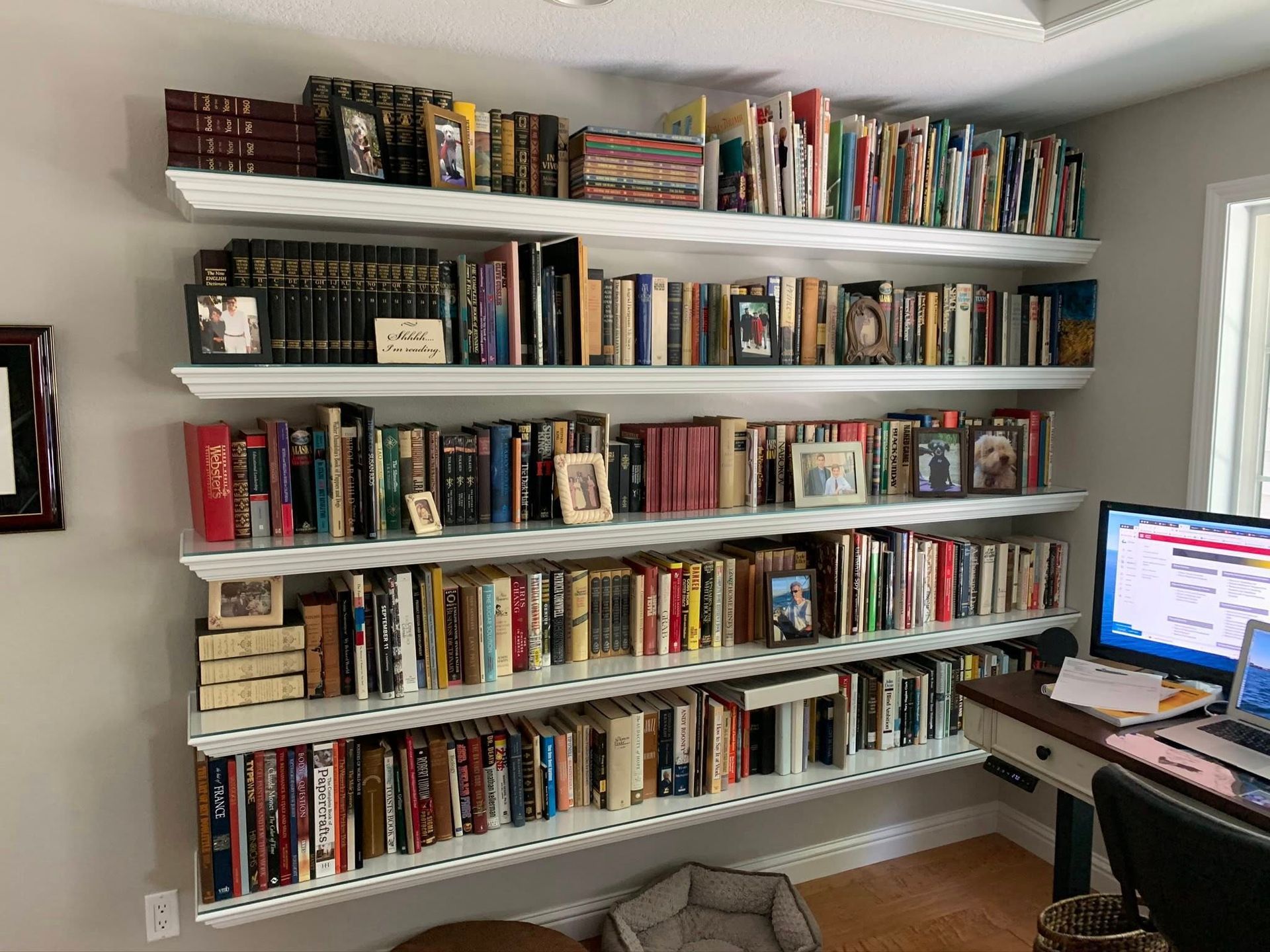 White floating bookshelves filled with books against a pale wall; a desk with a computer is to the right.