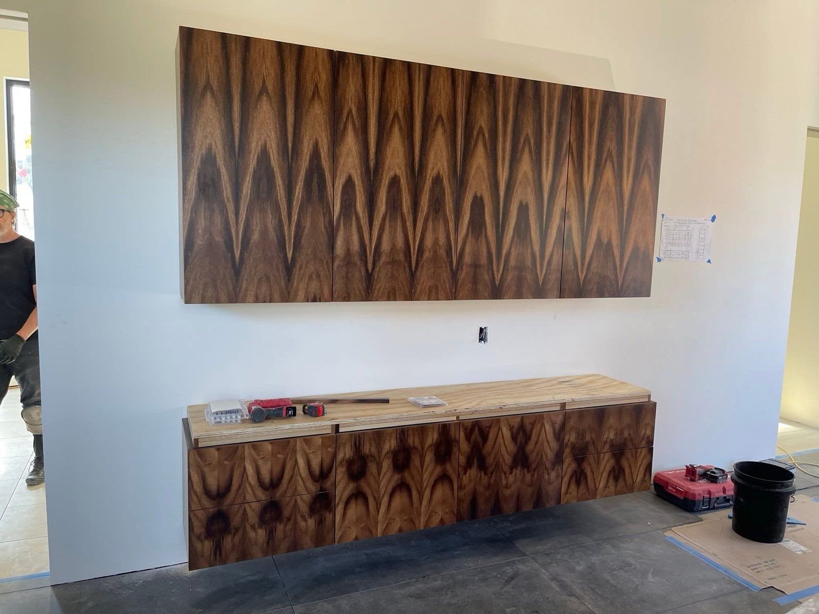 Wooden wall-mounted cabinets installed on a white wall, with a person standing nearby in a construction setting.