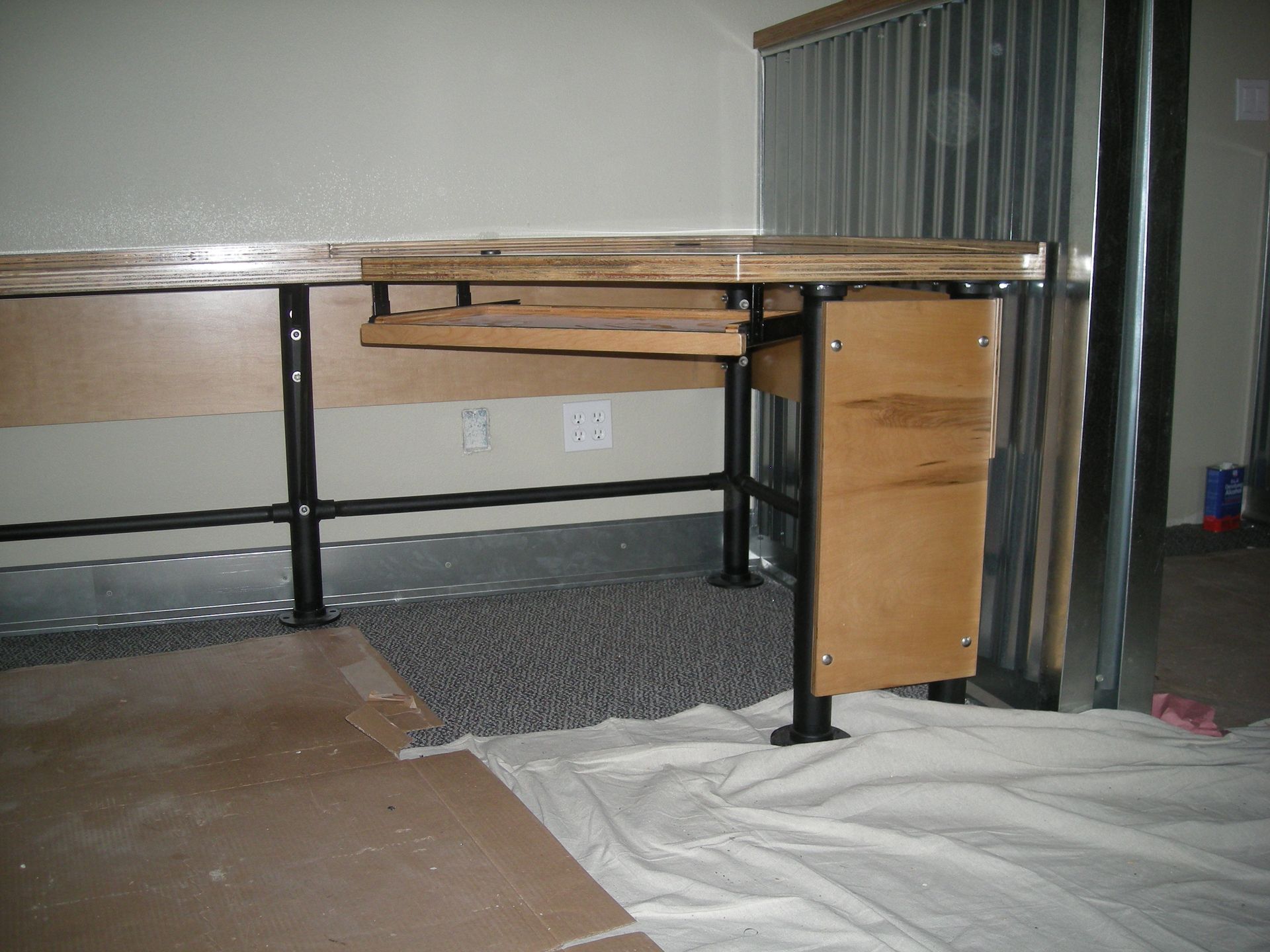A wooden desk with black metal legs is positioned against a wall with corrugated metal siding.