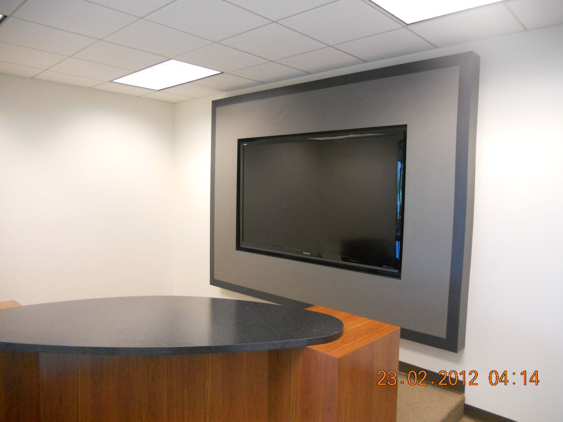 Large TV screen mounted on a wall with a gray border. A dark tabletop and wooden base are in the foreground.
