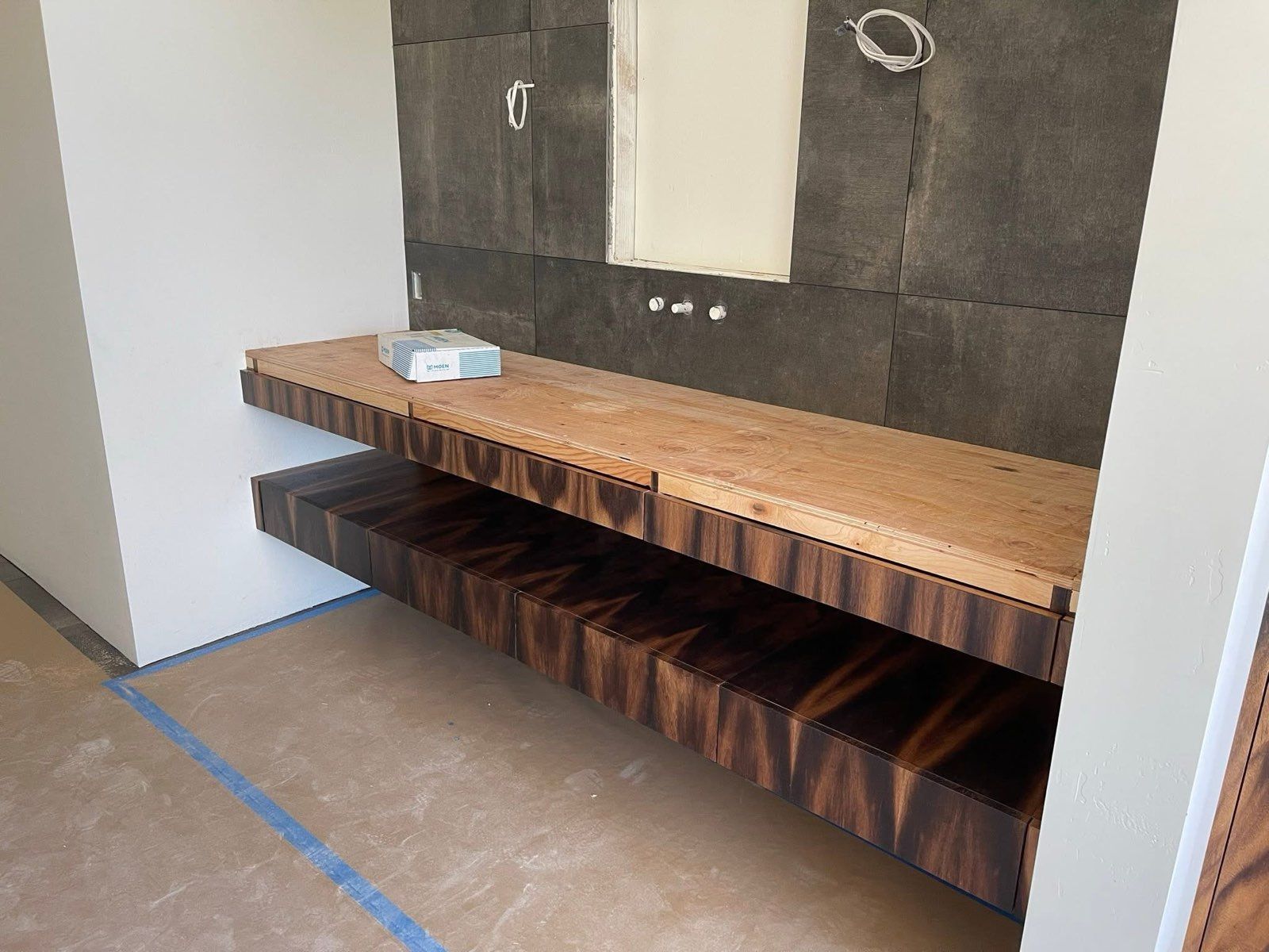 Floating wooden shelves in a bathroom under construction with dark tiled wall and wiring.