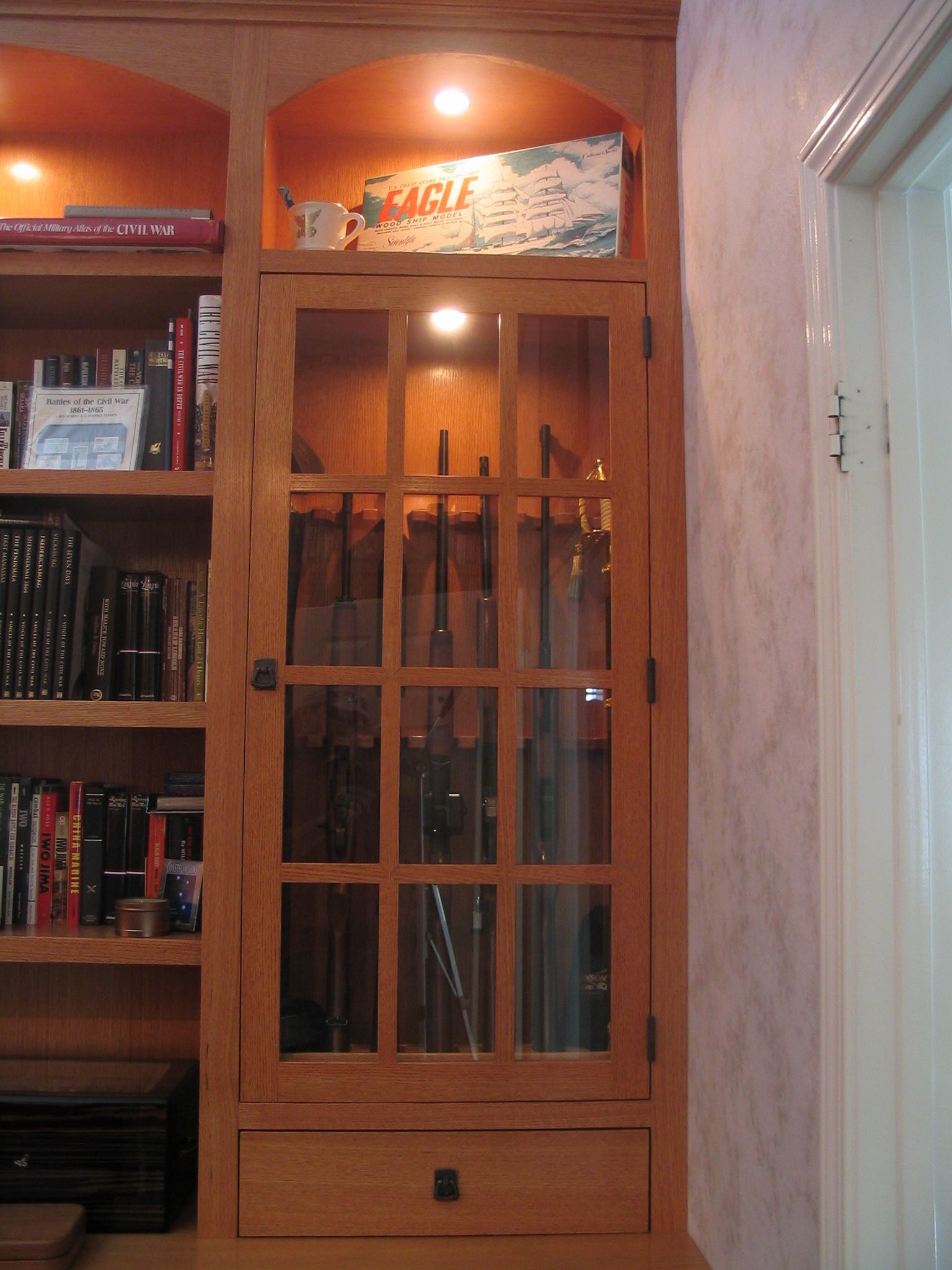 Wooden bookcase with glass-fronted cabinet containing rifles, lit from above, next to a door.