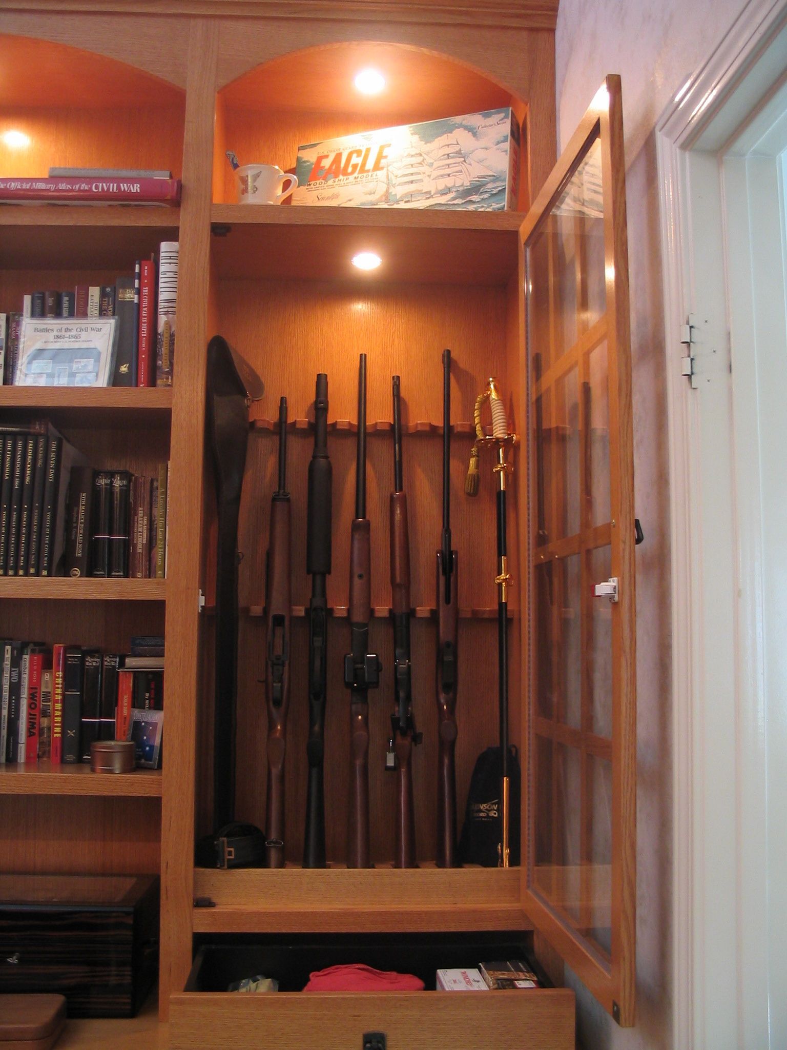 Wooden cabinet with rifles displayed inside, glass door open, built-in bookshelf.