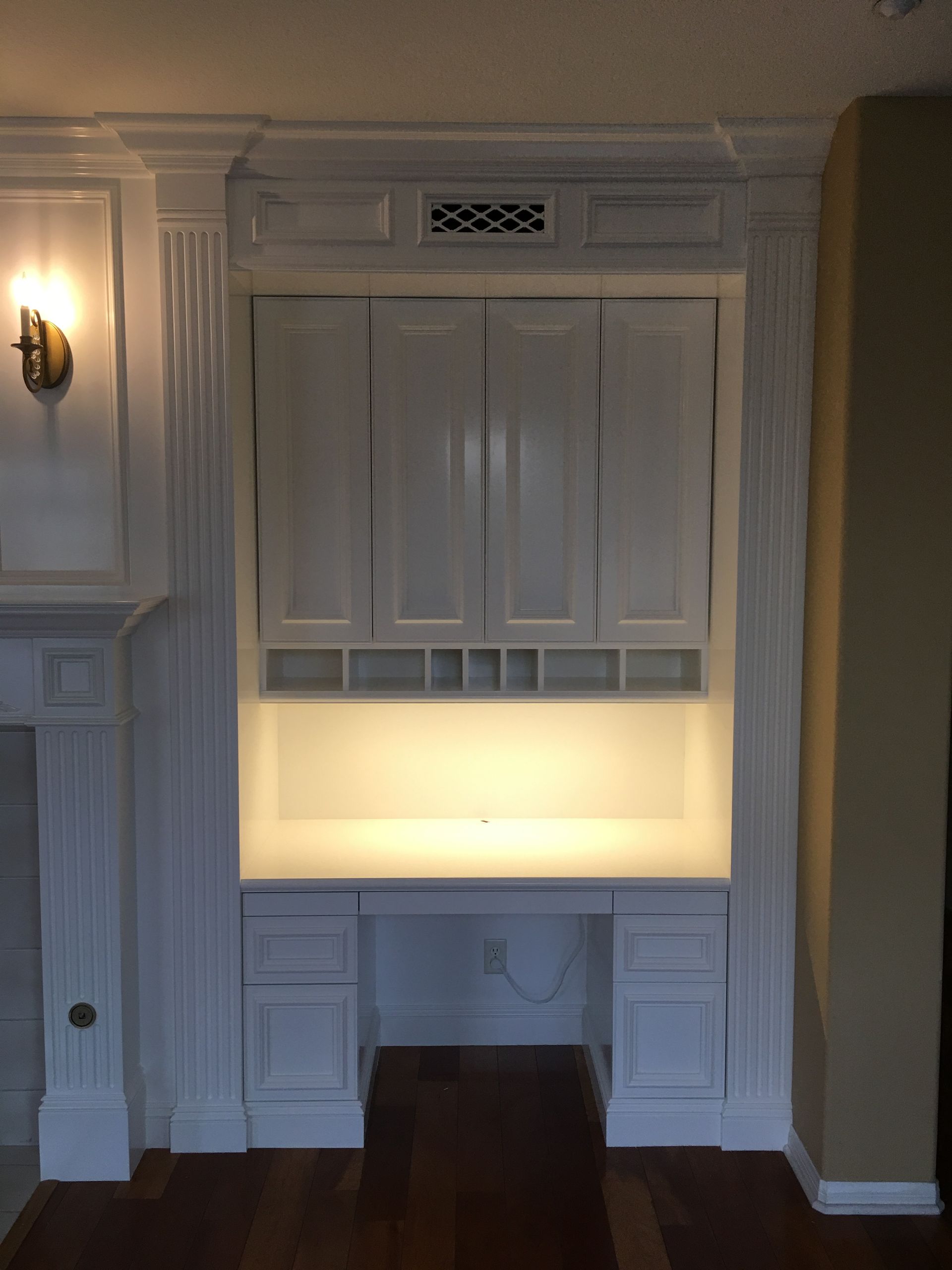 Built-in white desk with cabinets, overhead lighting, and storage cubbies, framed by white molding.