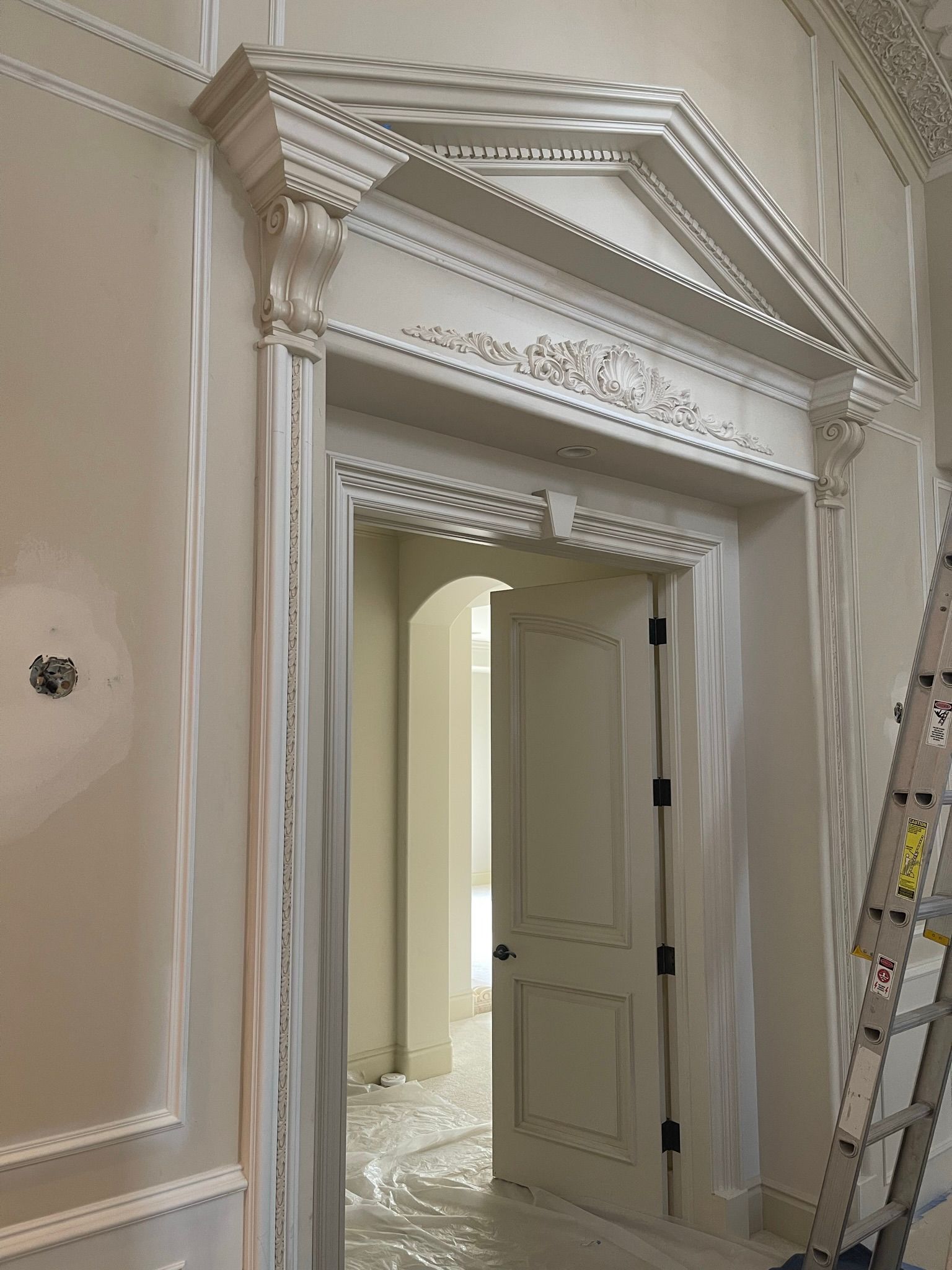 White doorway with ornate molding, ajar to reveal a light-filled room. A ladder leans against the wall.