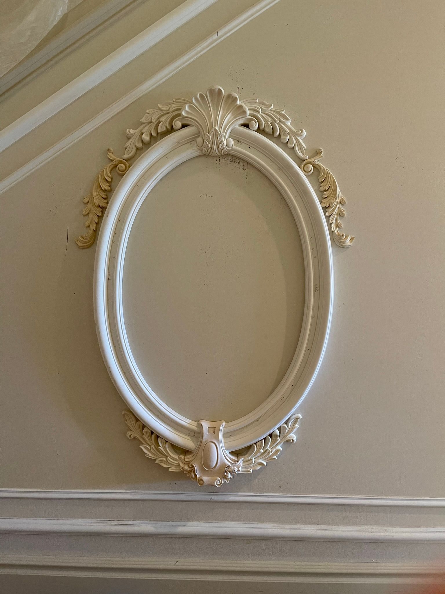 White oval decorative wall frame with ornate gold accents on a cream-colored wall.