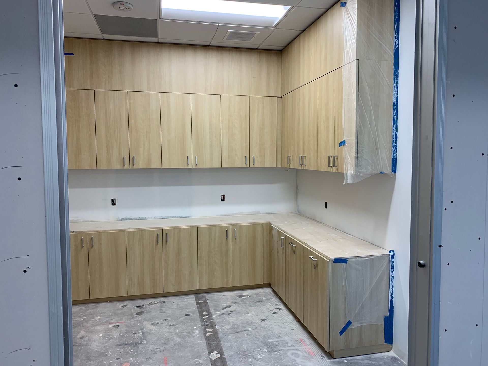 Room with light wood cabinets, upper and lower, along two walls, unfinished construction.