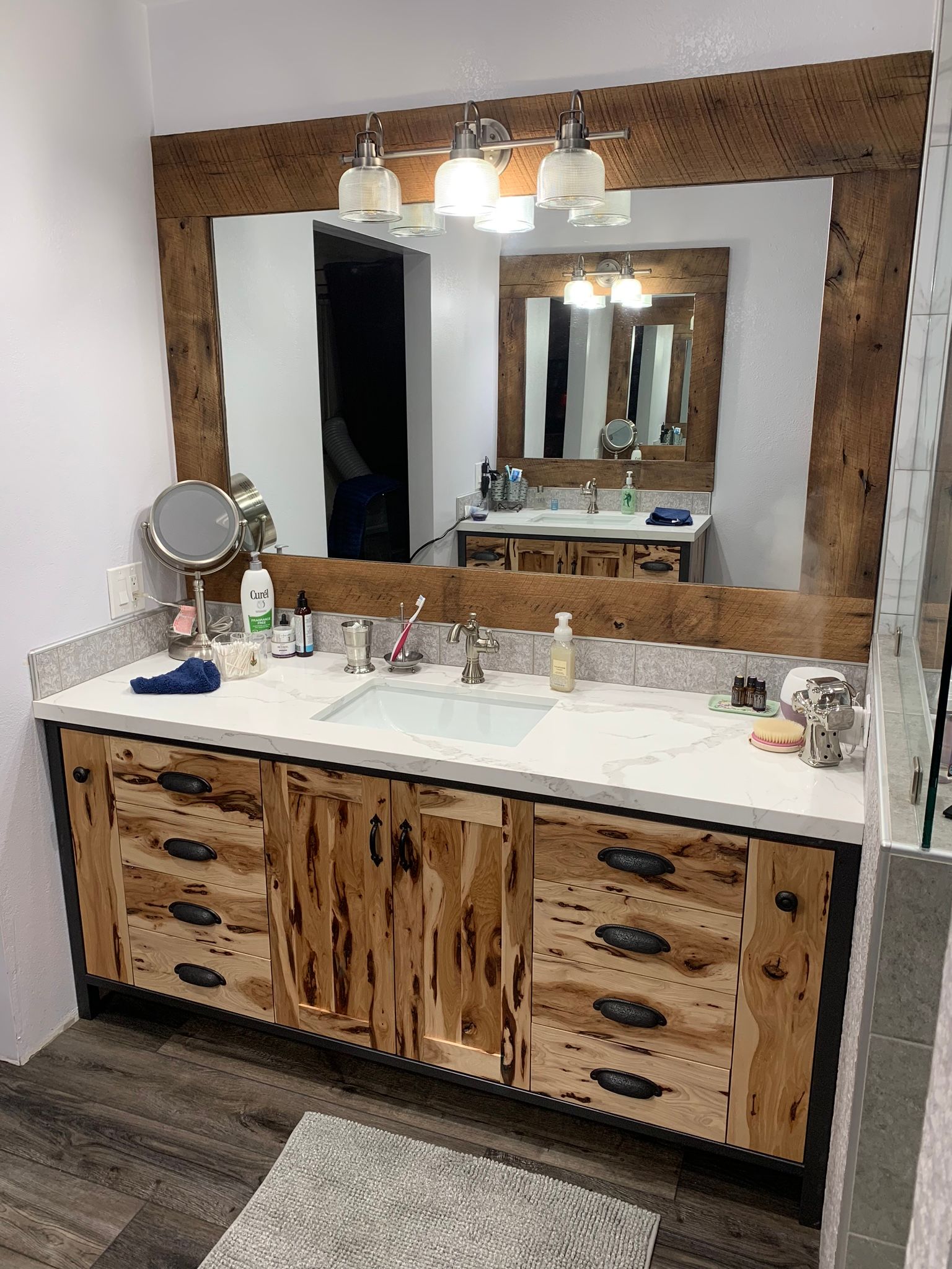 Rustic bathroom vanity with light wood cabinets, marble countertop, large mirror, and three-light fixture.