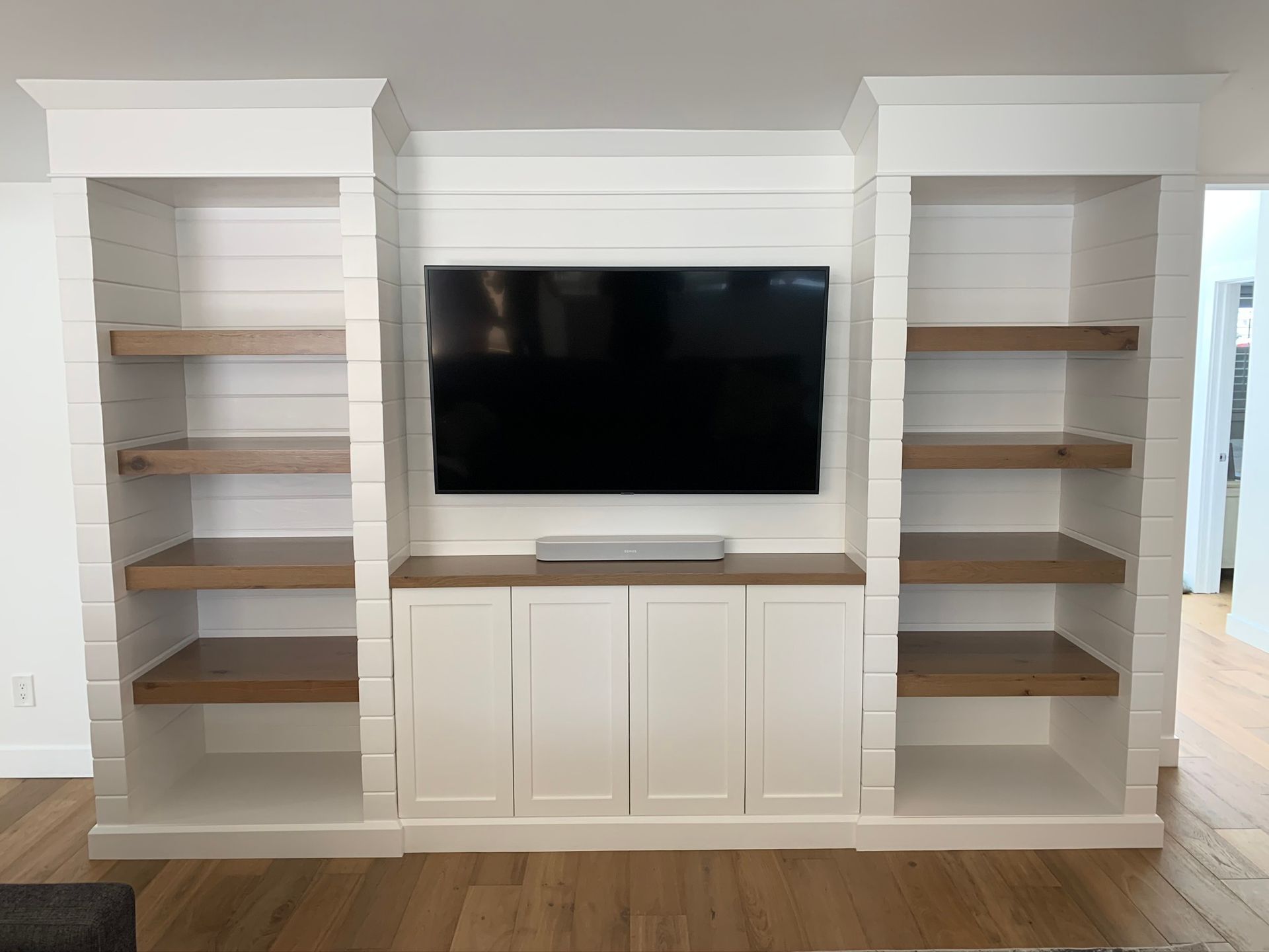 Built-in white media center with shelves, cabinets, and a mounted TV. Brown wooden shelves and a soundbar.