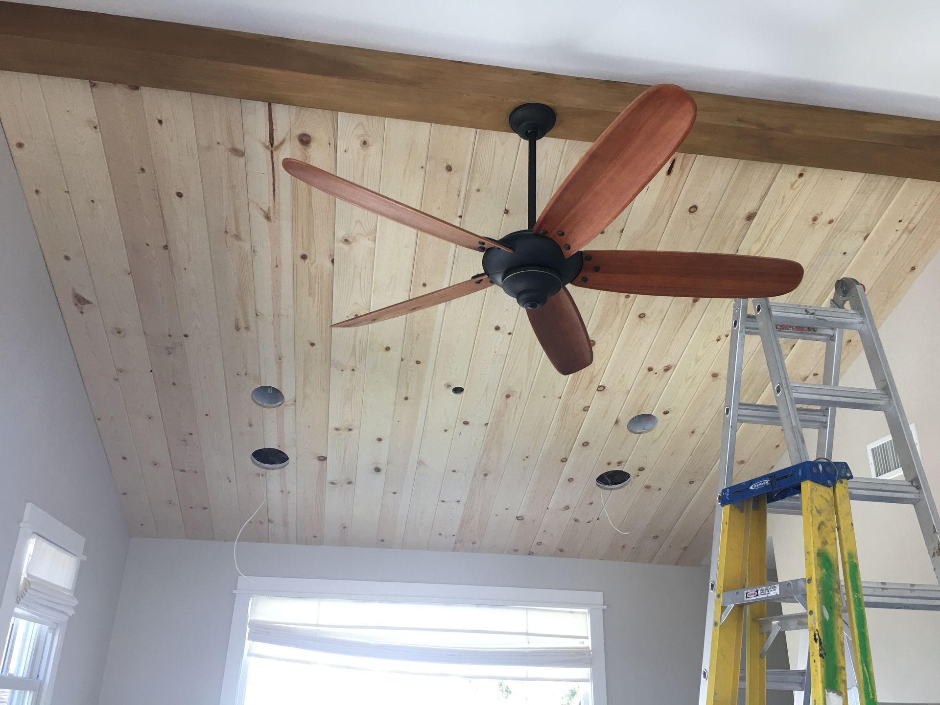 Ceiling fan with wooden blades installed on a wood plank ceiling, ladder in foreground.
