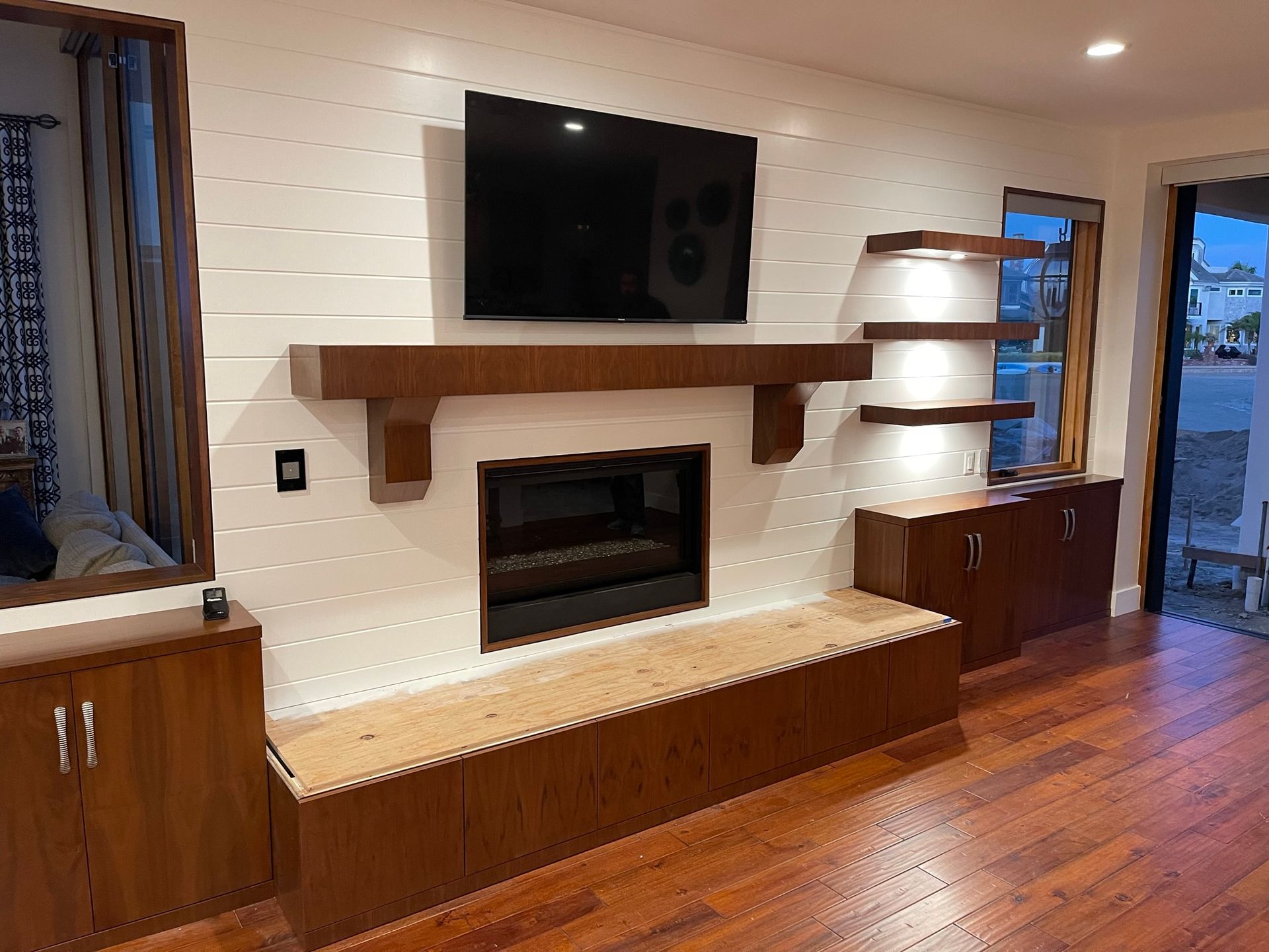 Living room with fireplace, TV, and shelves on a white shiplap wall. Wooden cabinets and flooring.