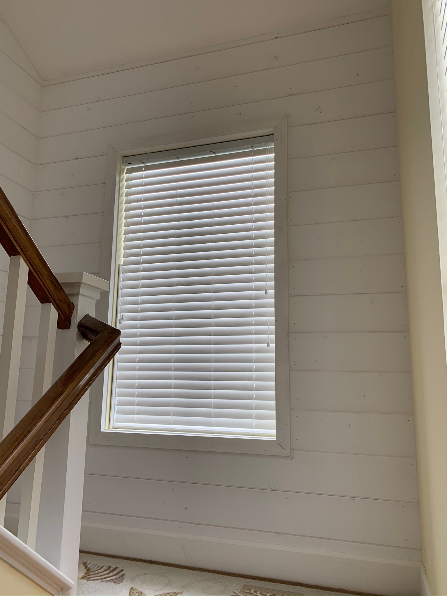 Window with closed blinds on a white-planked wall; stairs with wooden banister in the corner.