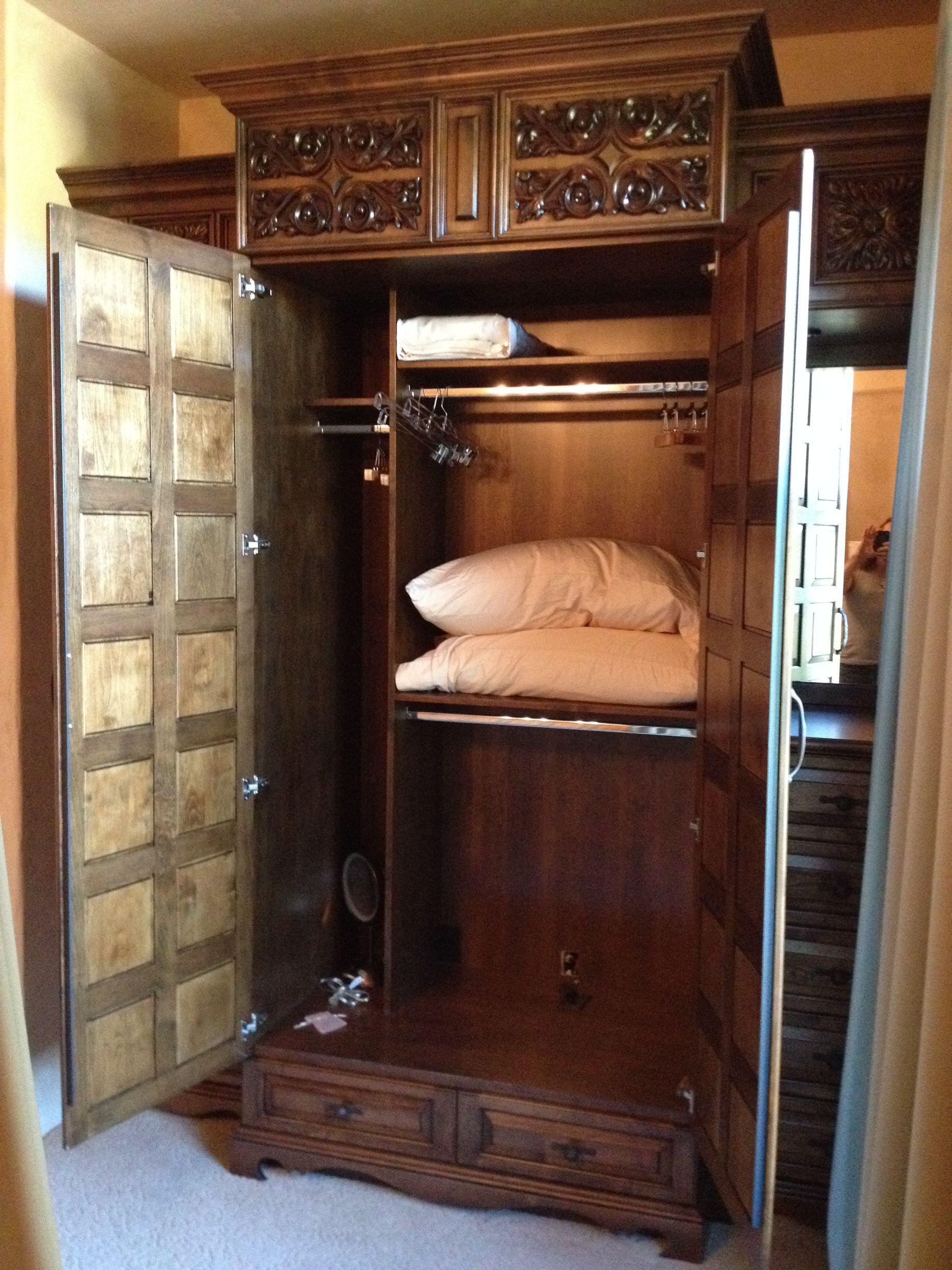 Wooden wardrobe with carved details, open to reveal storage, pillows, and a hanging rod.
