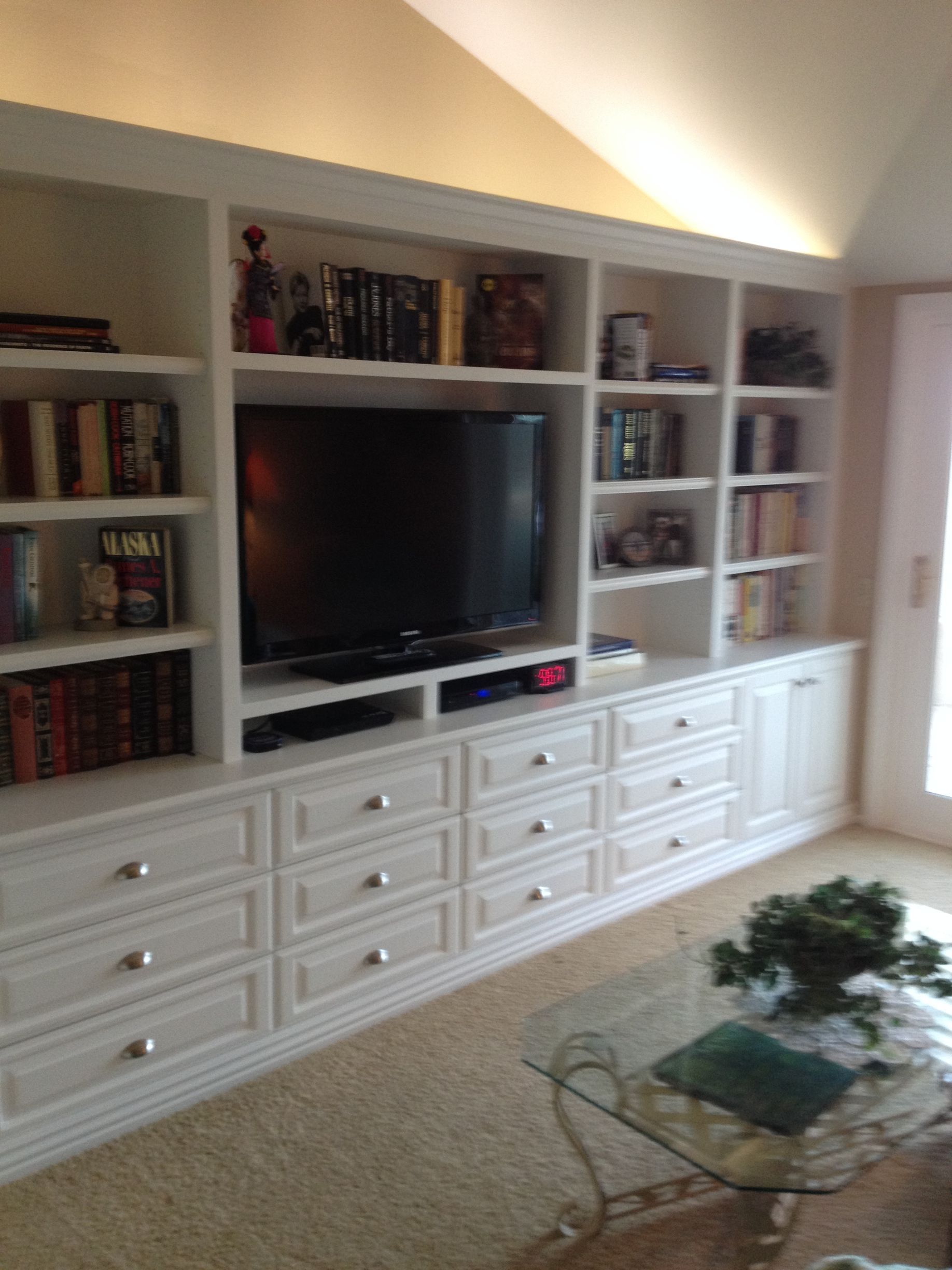White built-in entertainment center with drawers, shelves, and a TV. Books and decorative items are displayed.
