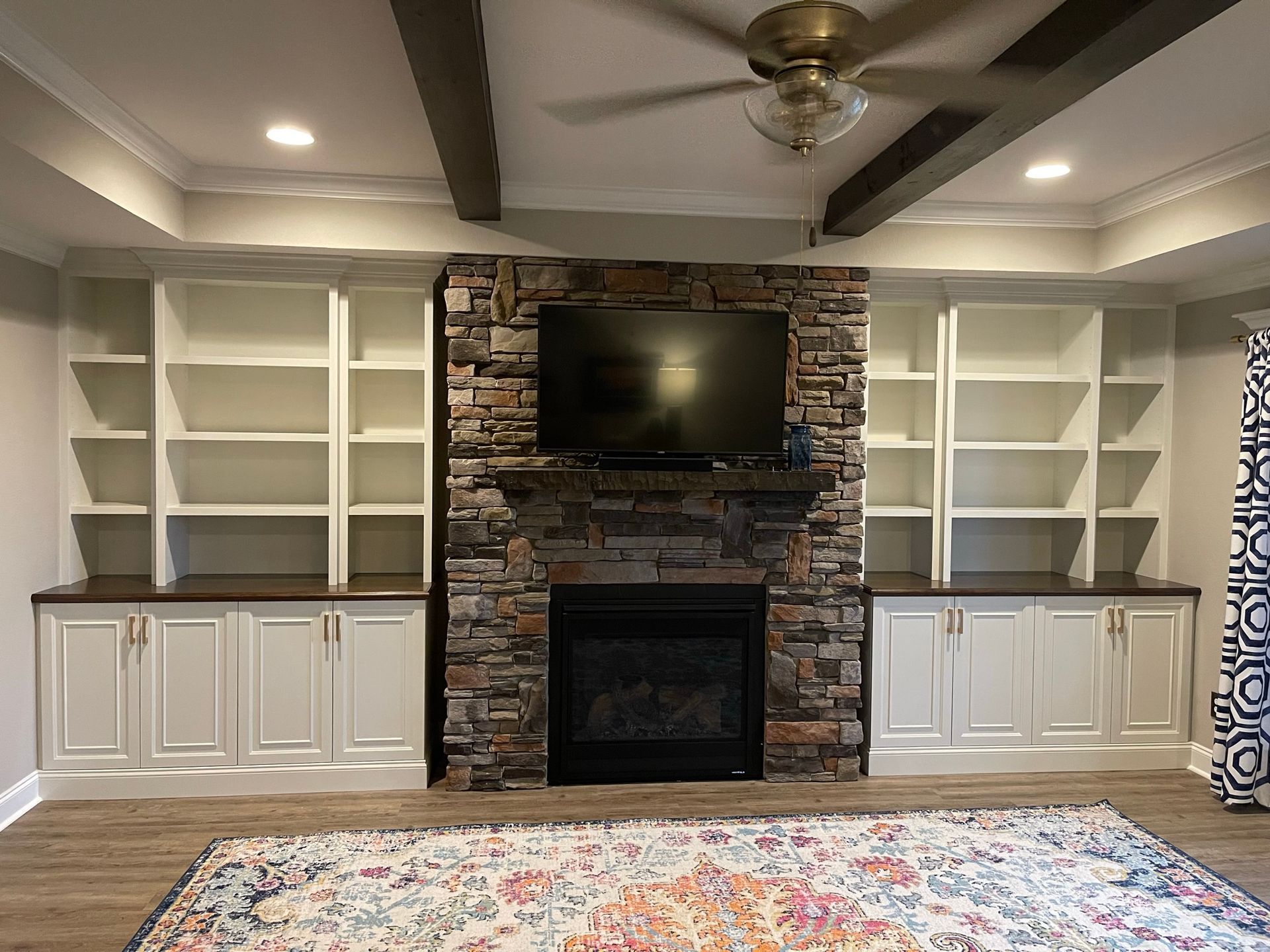 Fireplace with stone surround, flanked by white bookshelves. TV above, ceiling beams, and rug in front.