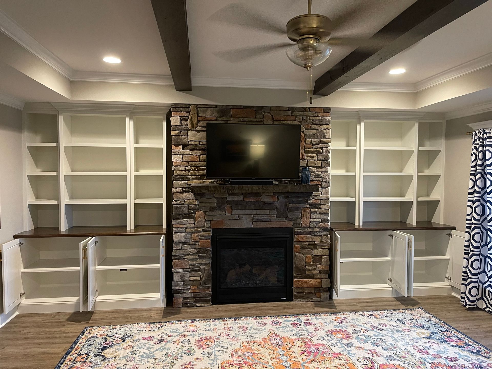 Built-in bookshelves flank a stone fireplace with a TV above. Dark ceiling beams, white shelves, and a patterned rug.