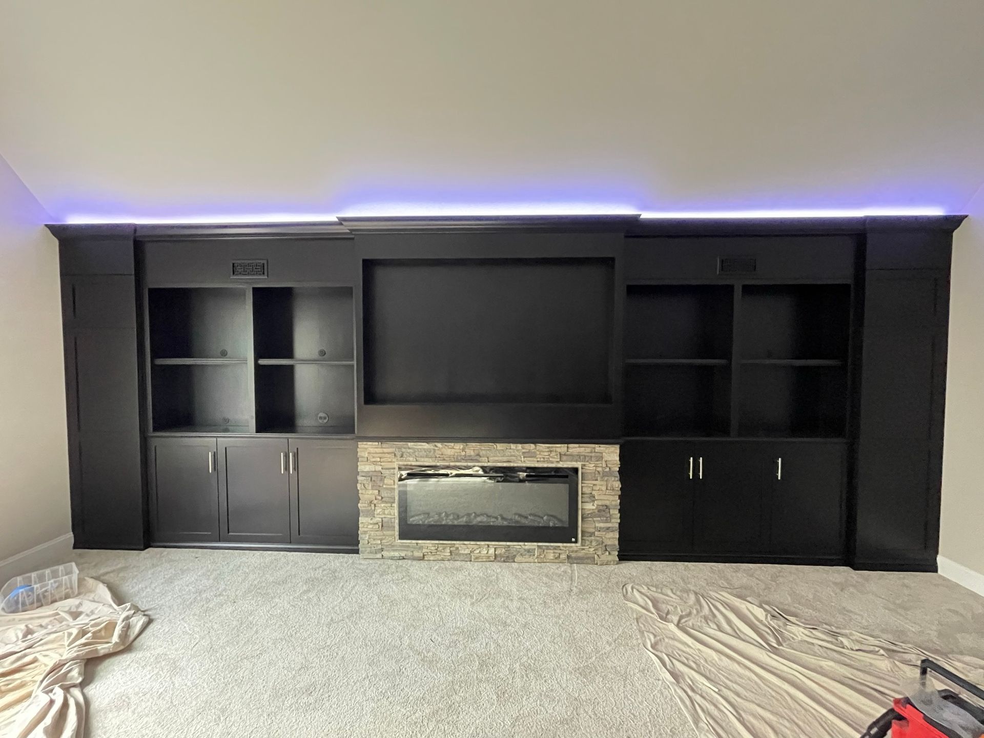 Black media center with a fireplace and shelves, set against a grey wall and carpet; LED lighting along the top.