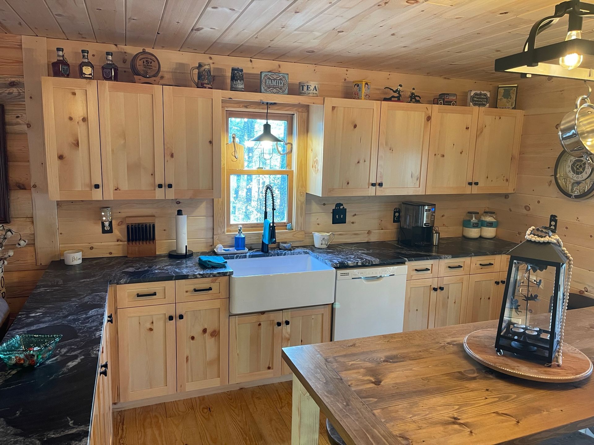 Rustic cabin kitchen with light wood cabinets, dark countertops, farmhouse sink, and a window overlooking trees.
