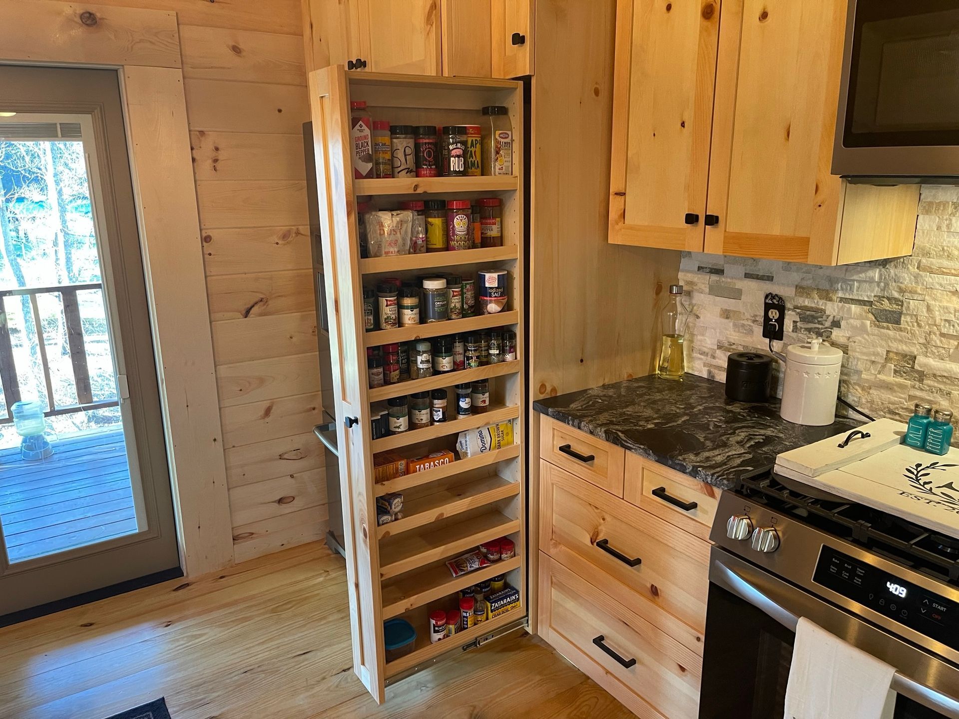 Wooden kitchen with spice rack pull-out. Open door reveals spices. Cabin-style interior, wooden cabinets.