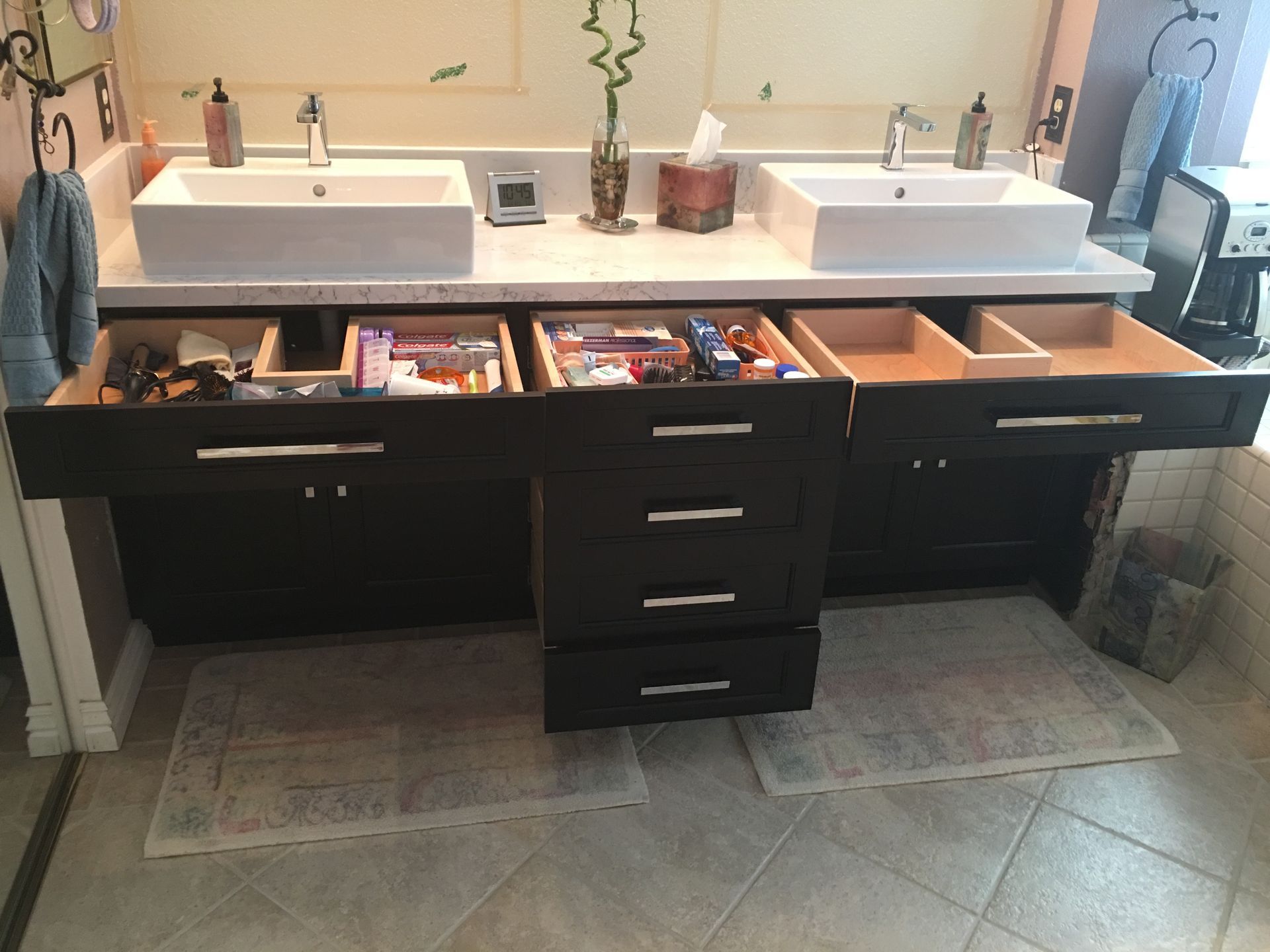 Bathroom vanity with open drawers, two sinks, and toiletries. Brown cabinets, white countertops, and gray floor mats.