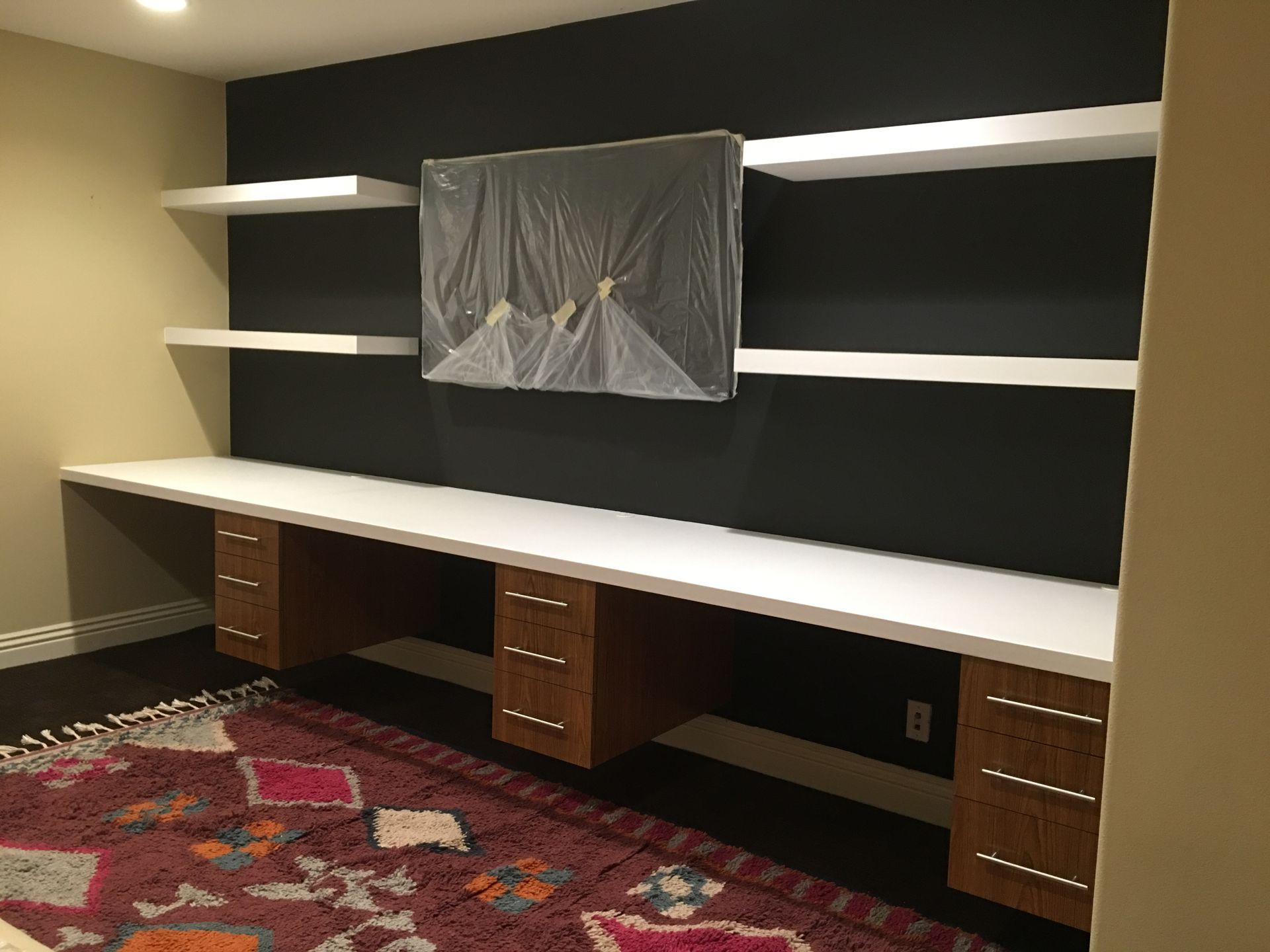Modern office space with long white desk, floating shelves, and TV on black wall.