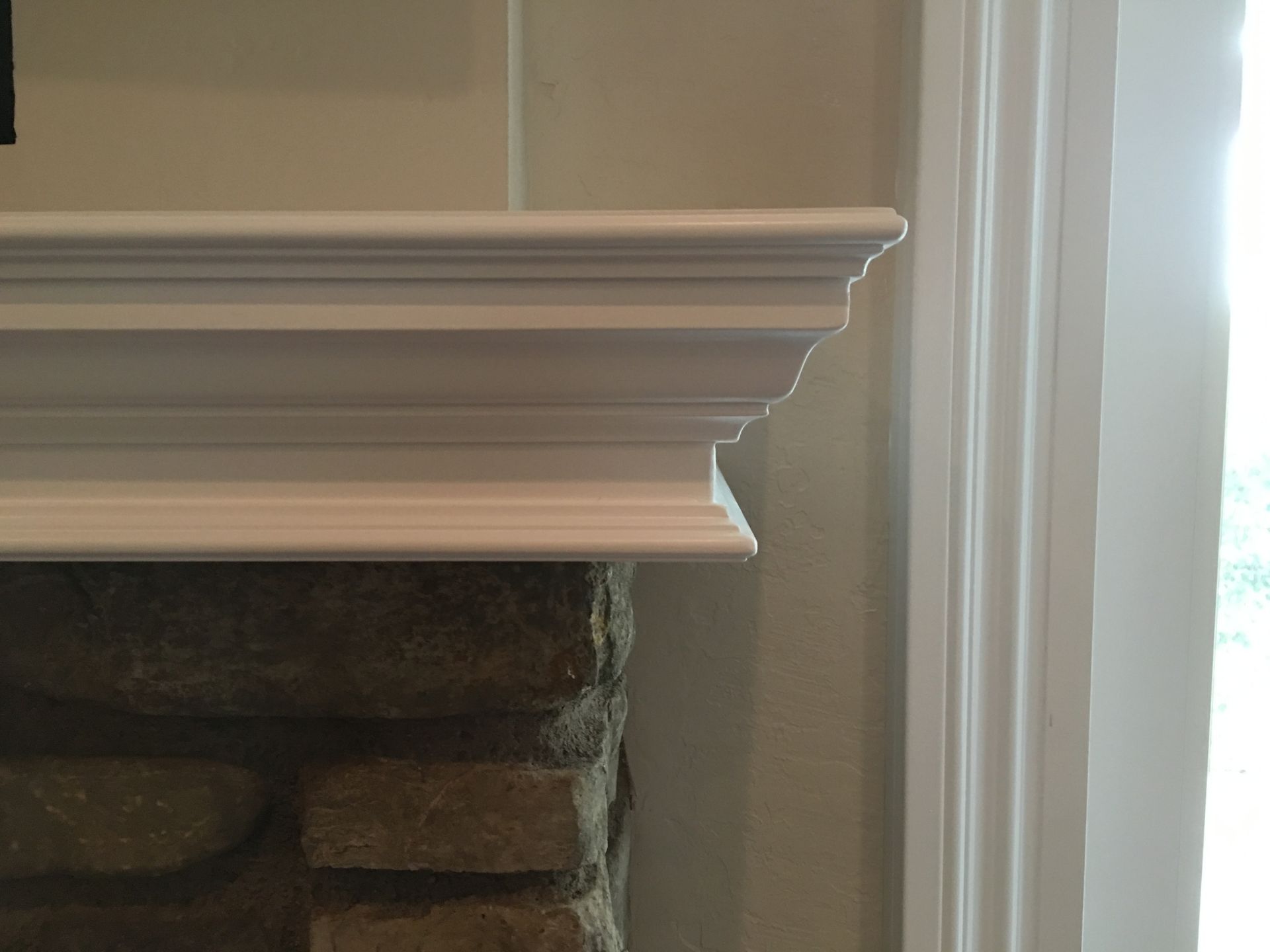 White mantelpiece on a stone fireplace next to a window frame.