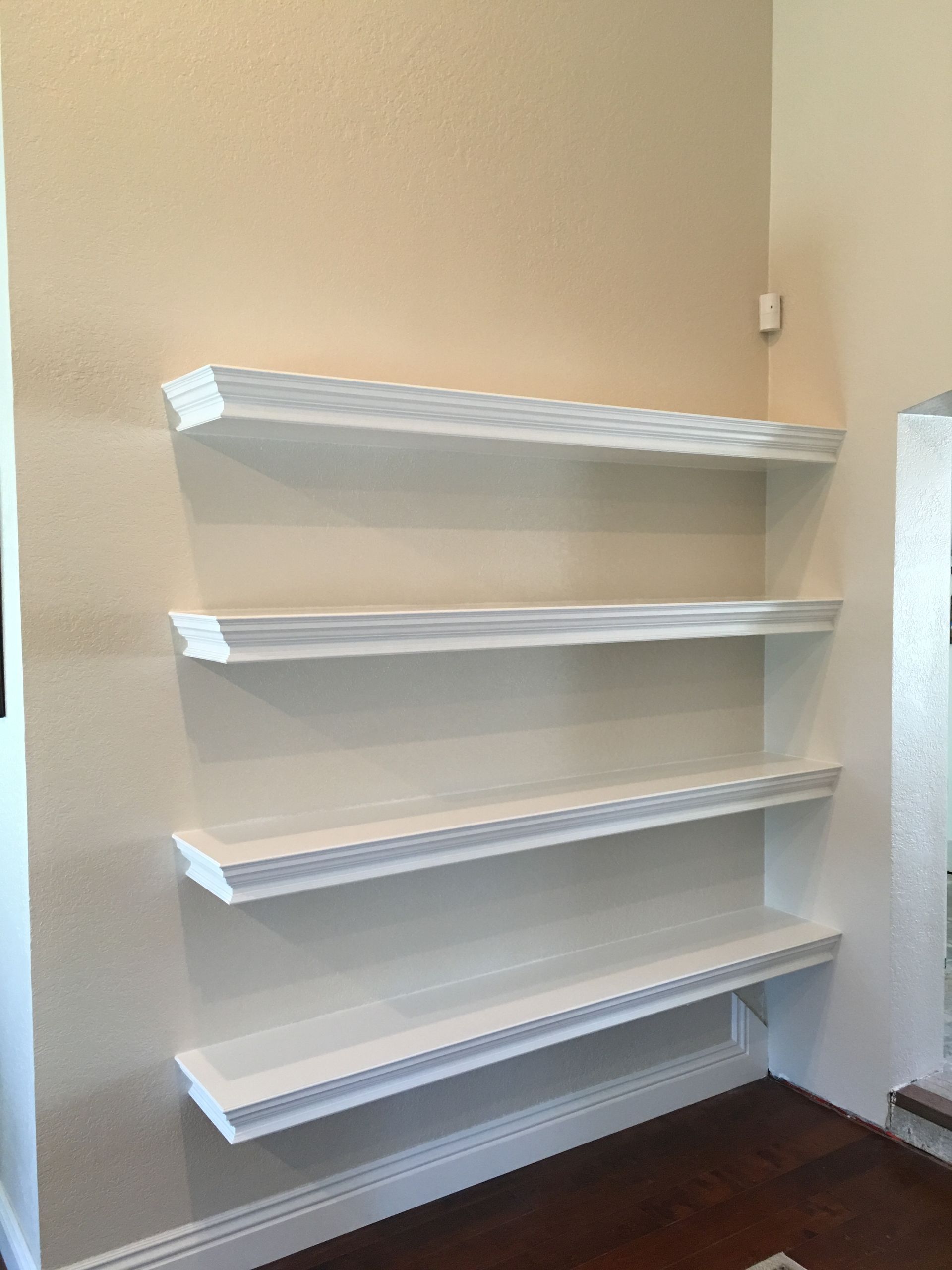Three white shelves on a beige wall, wooden floor, corner setting.