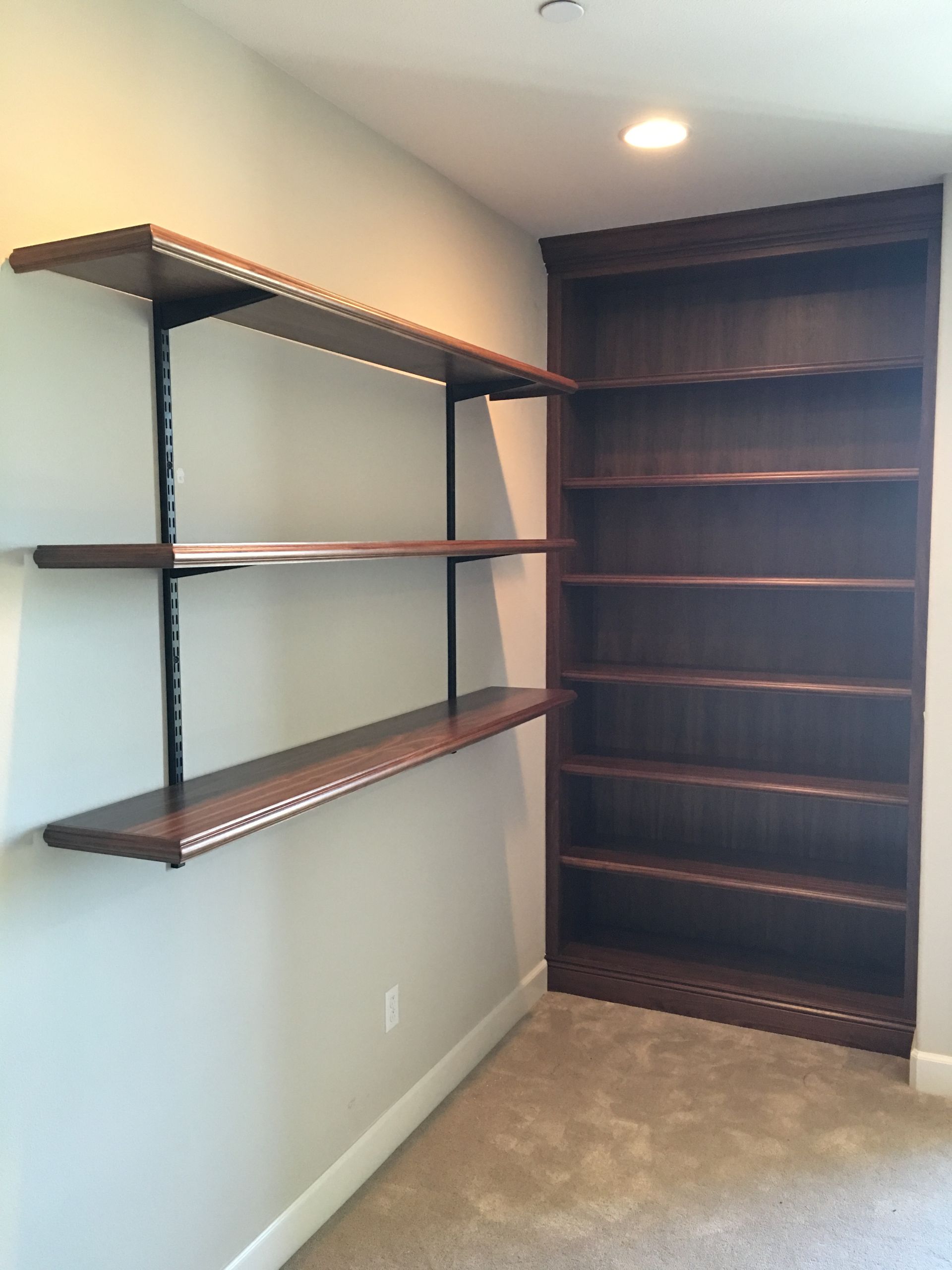 Built-in dark wood bookshelves in a corner. Floating shelves on the left. Beige walls, carpeted floor.