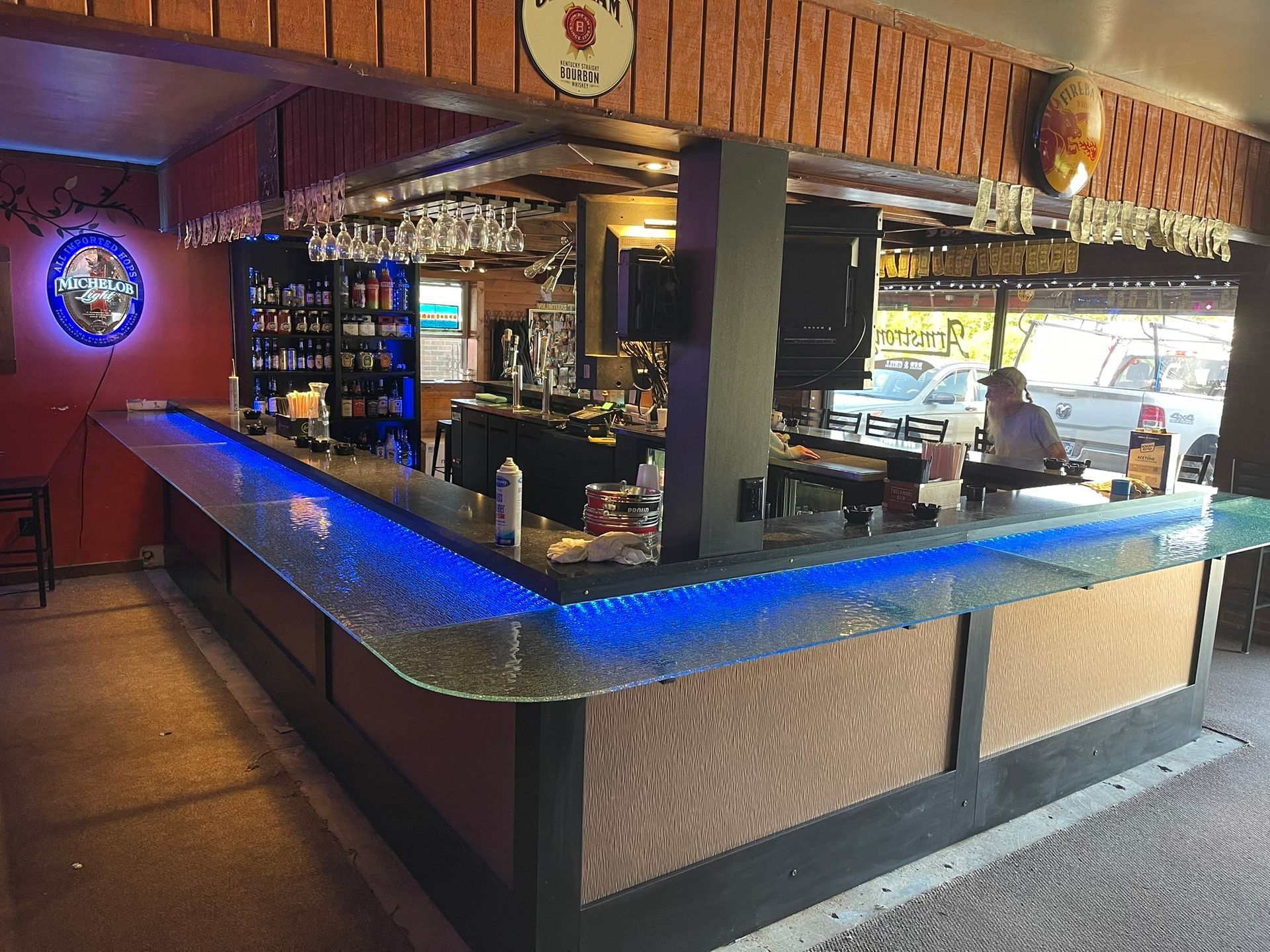Bar with blue-lit countertop, liquor bottles, and a person behind the counter. Outside light visible.