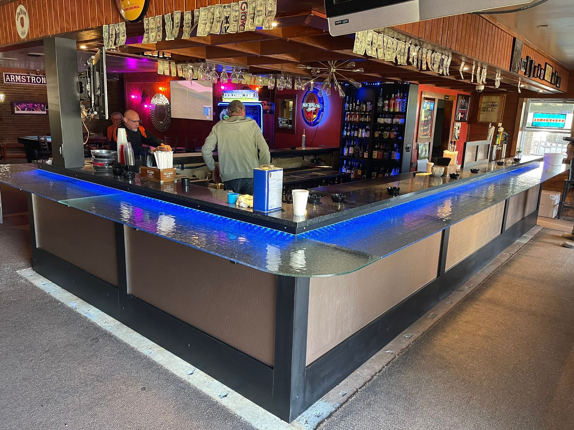 Bar with blue-lit countertop, bartenders, shelves of alcohol, and a few patrons.