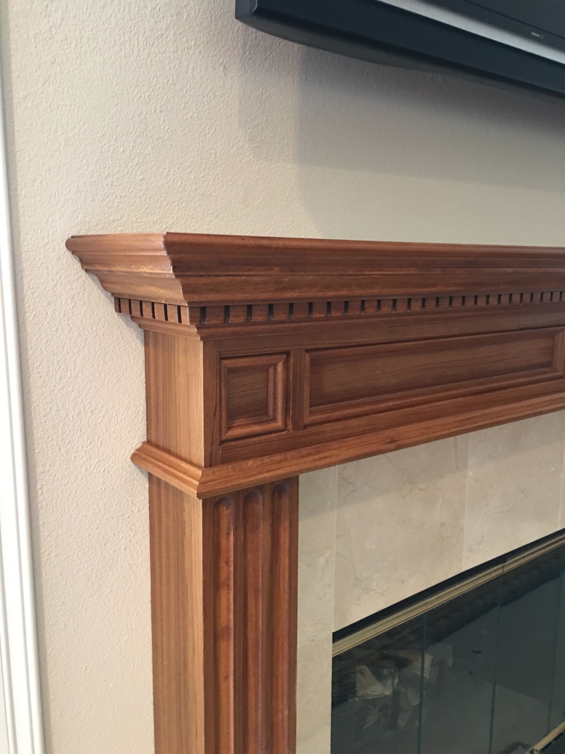 Wooden fireplace mantel with ornate details, against beige wall, fireplace visible below.