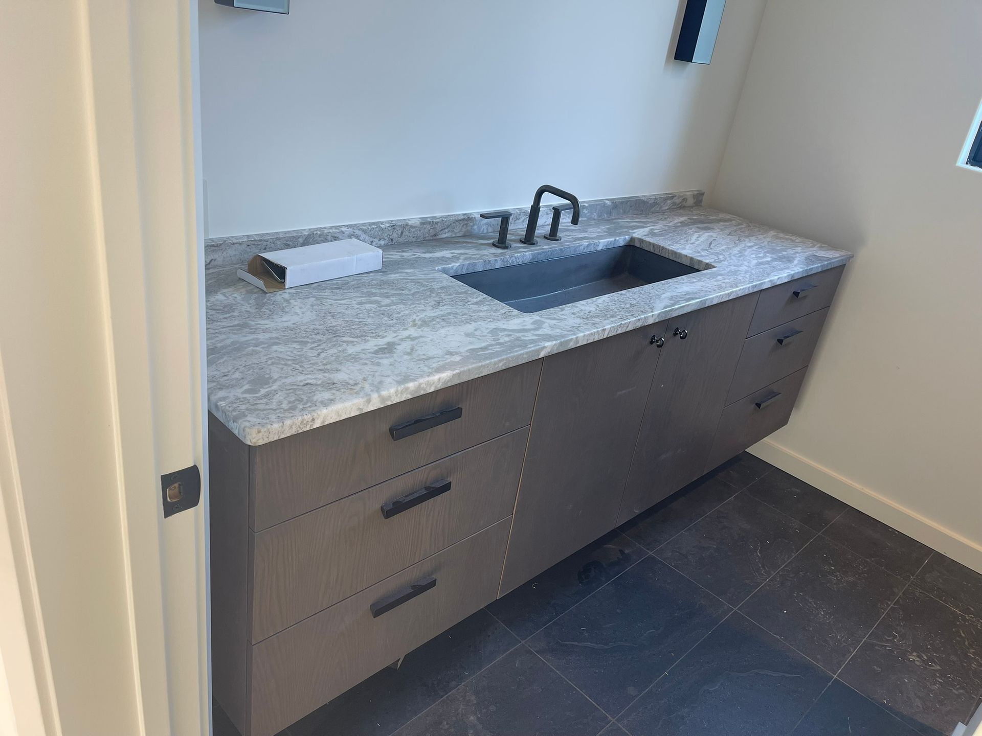 Gray floating vanity with black faucet and sink, granite countertop, and three drawers in bathroom.