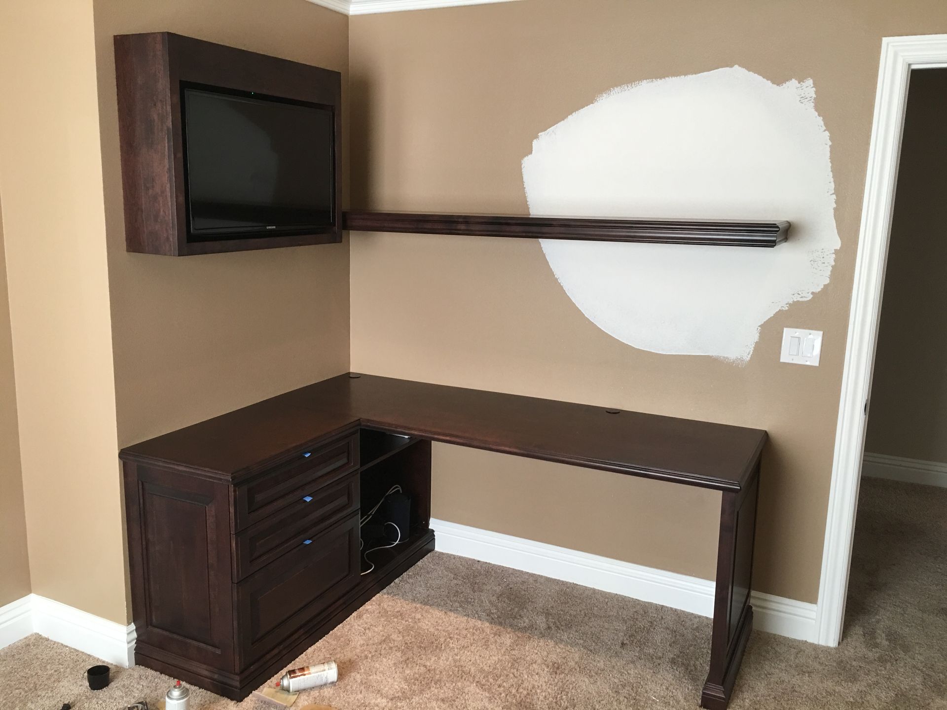 L-shaped dark wood desk with attached cabinet and floating shelf; TV in wood box above. Tan walls.