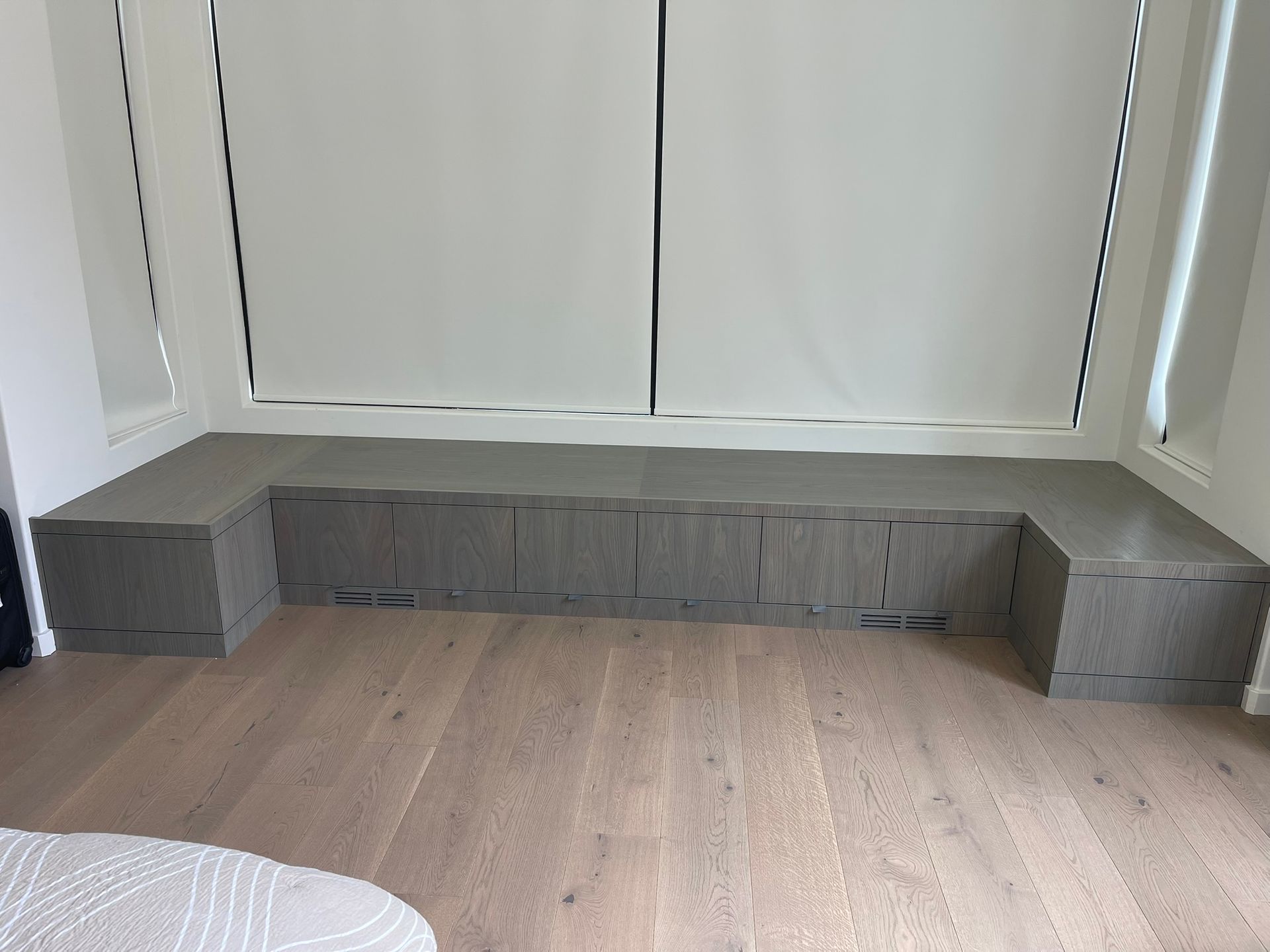 Built-in gray storage unit beneath a window with white blinds, set against light wood flooring.