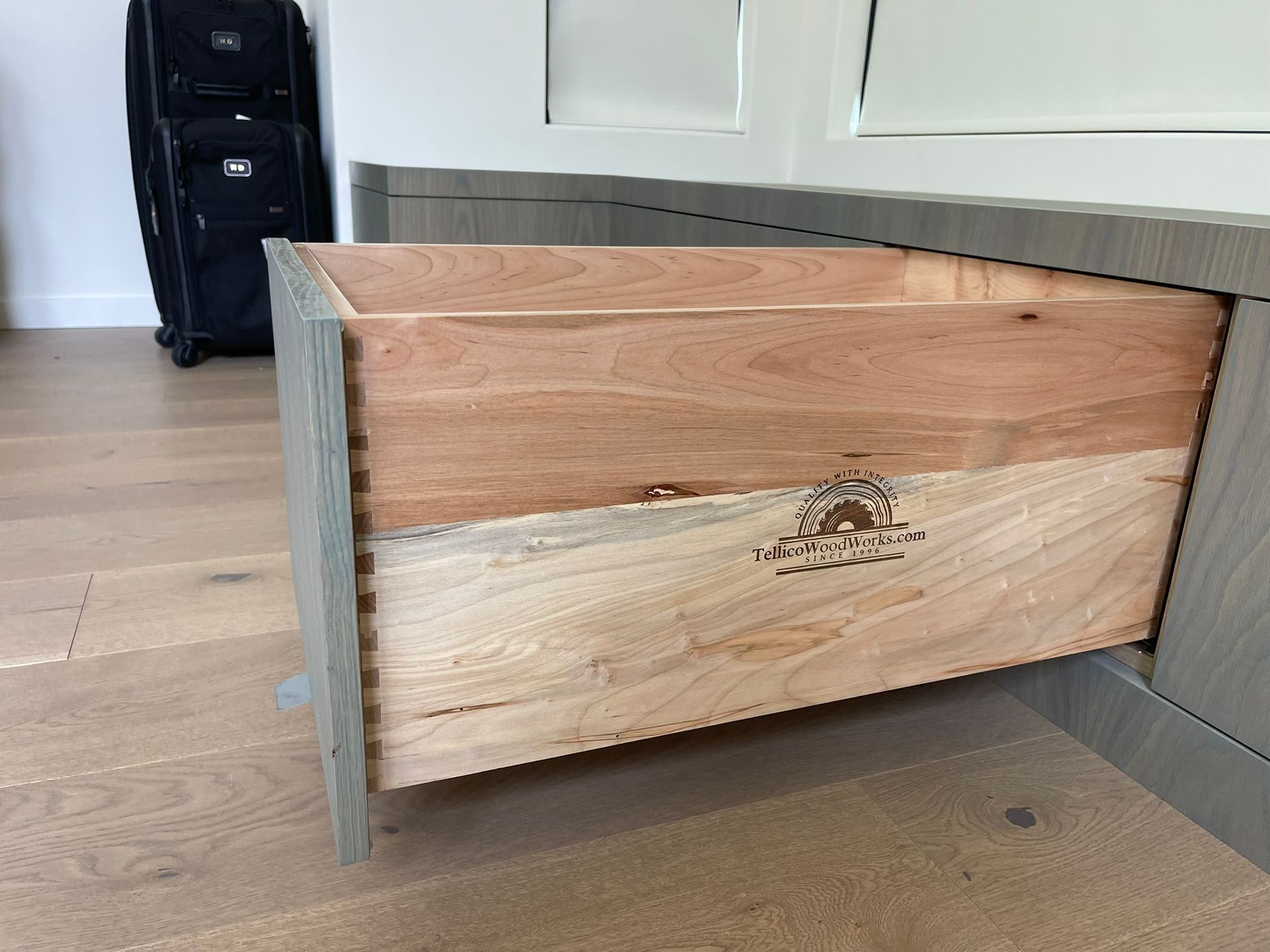 Open wooden drawer in a gray cabinet, showing dovetail joints and a branded stamp.