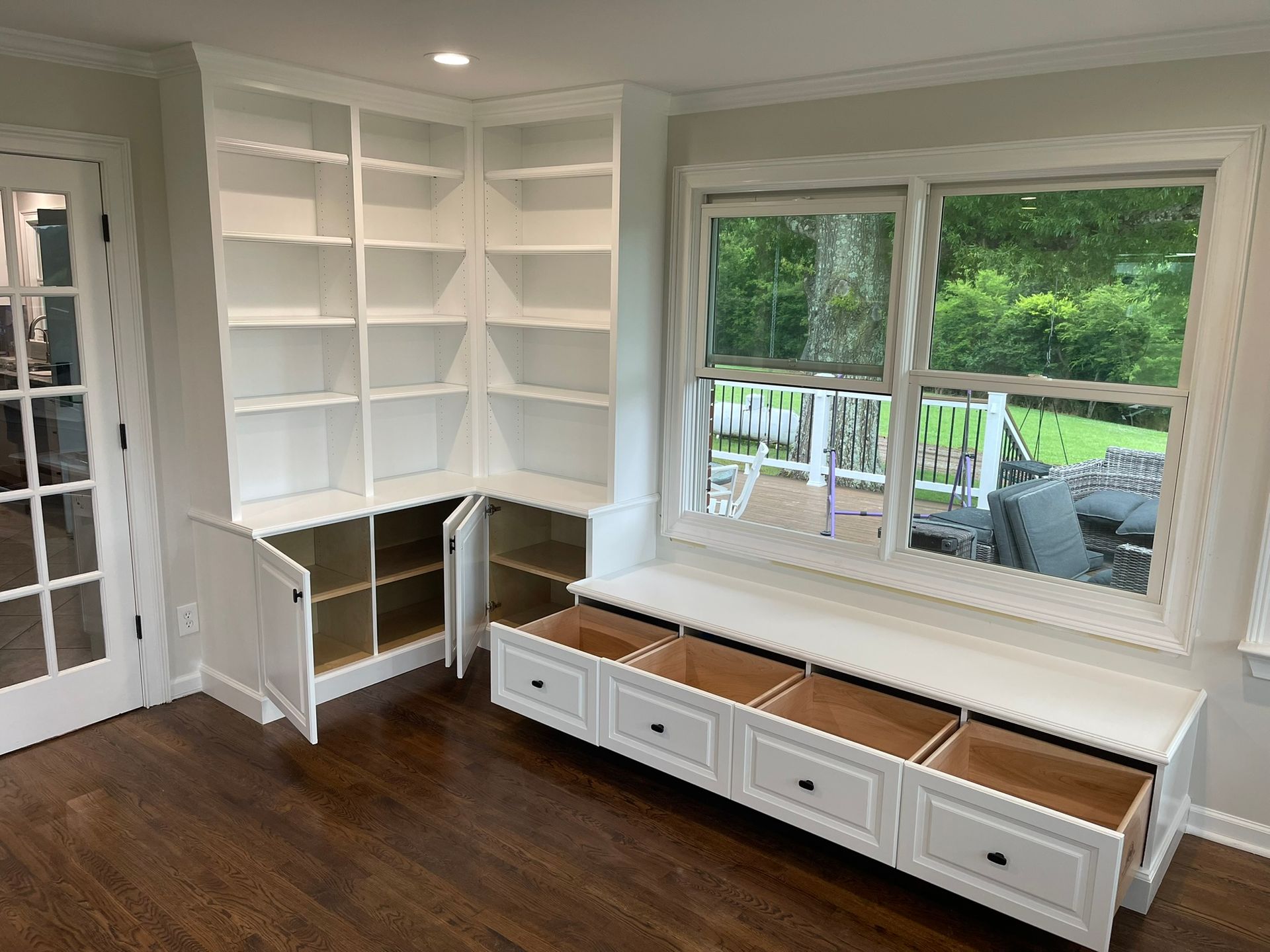 White built-in shelves and window seat with open drawers and cabinet doors against a window overlooking a deck.