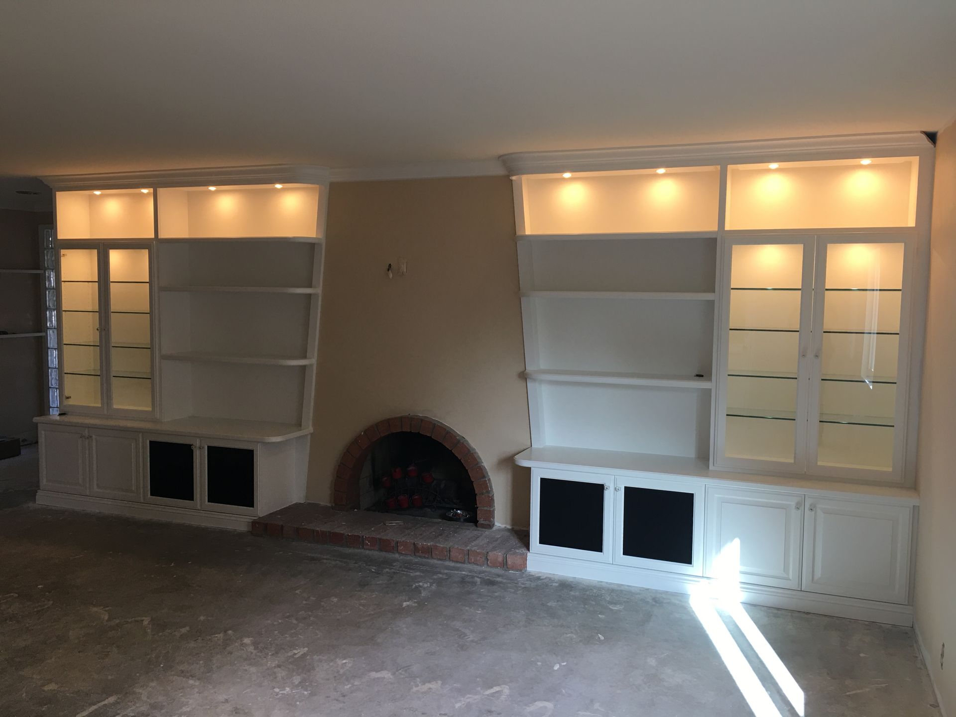 White built-in bookshelves with glass doors flank a brick fireplace. Shelves are lit with overhead lights, on light tan wall.