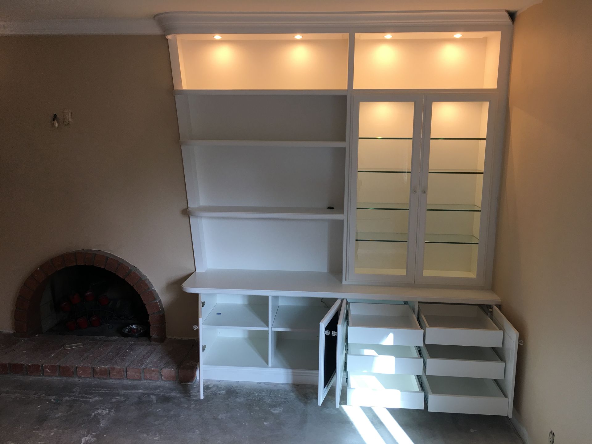 White built-in cabinet with glass doors, shelves, and drawers, illuminated by overhead lights, next to a fireplace.