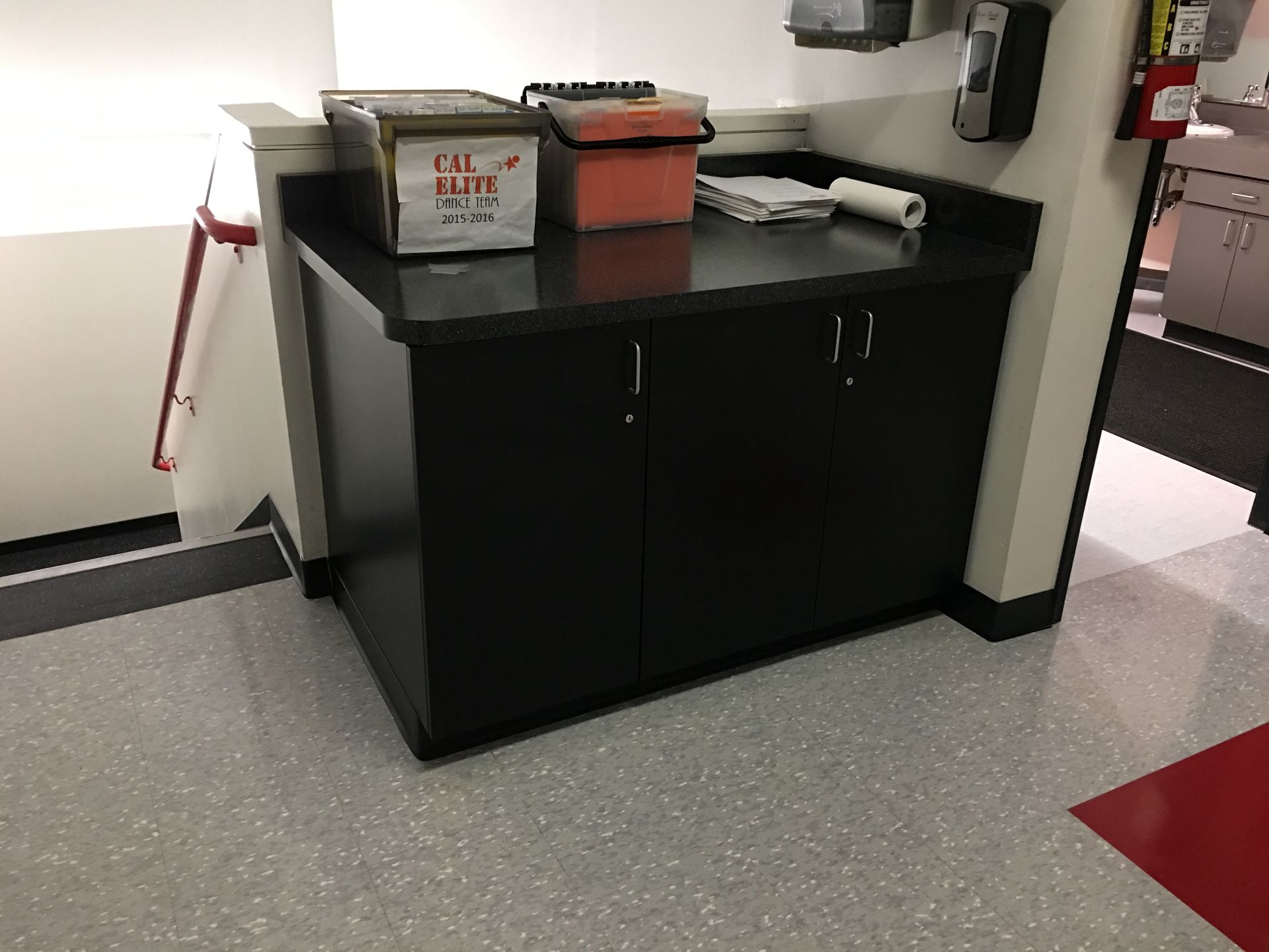 Black cabinet with supplies on countertop in a medical facility, next to a hallway.