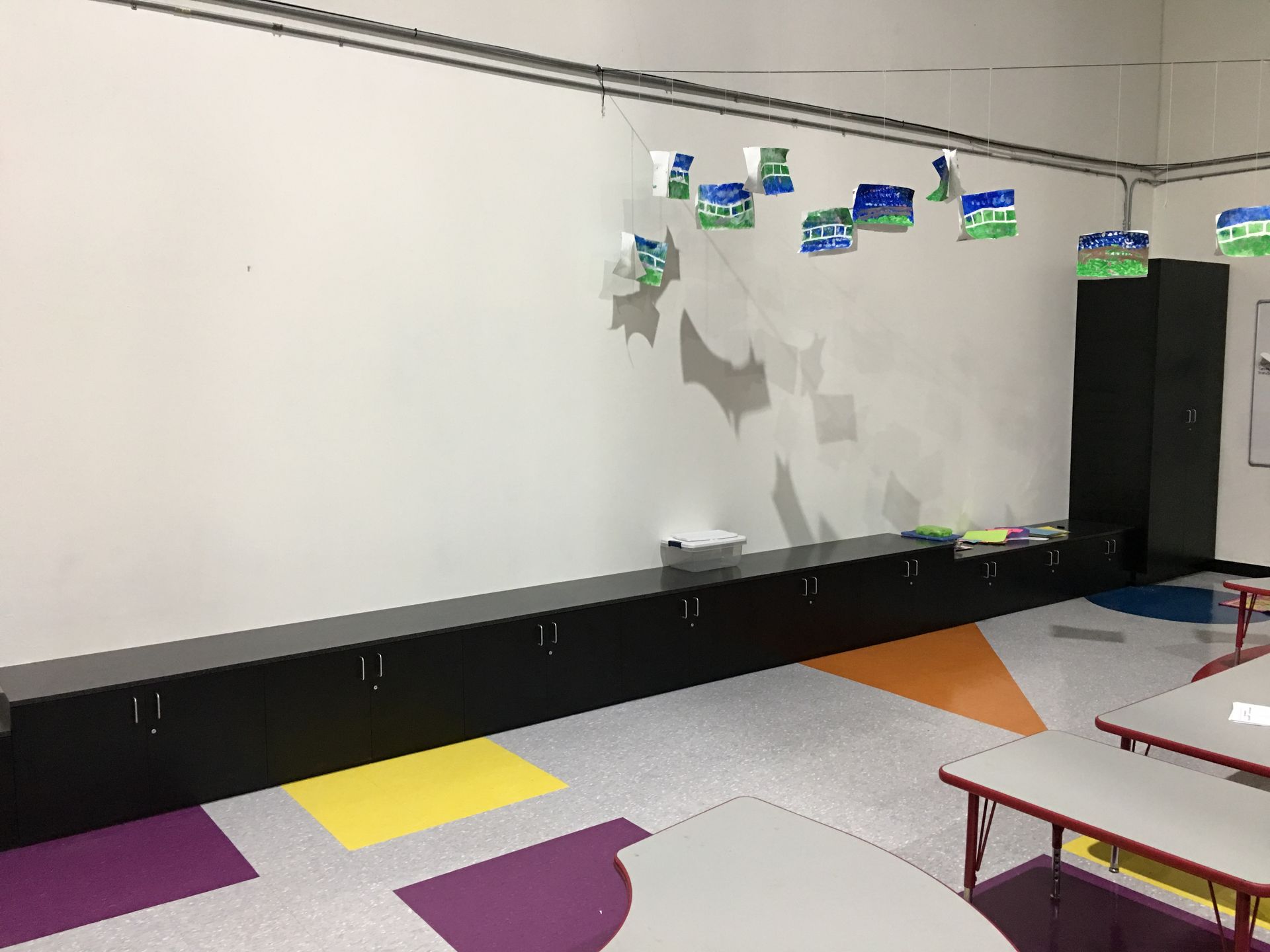 A classroom with a white wall, long black storage, colorful floor, and hanging decorations.